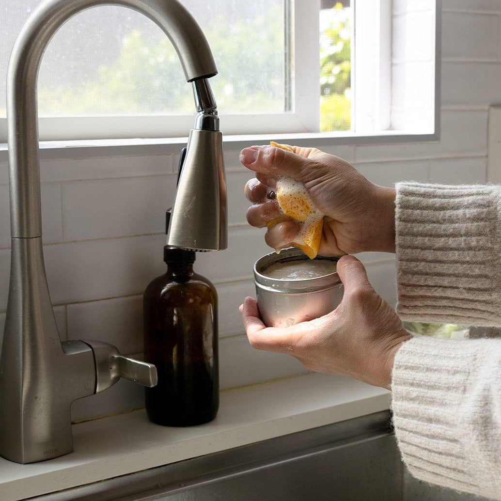 A person in a cream sweater squeezes a ZeroWasteStore.com Cellulose Kitchen Sponge over a round metal dish by the sink, with a brown bottle on the countertop near a window.