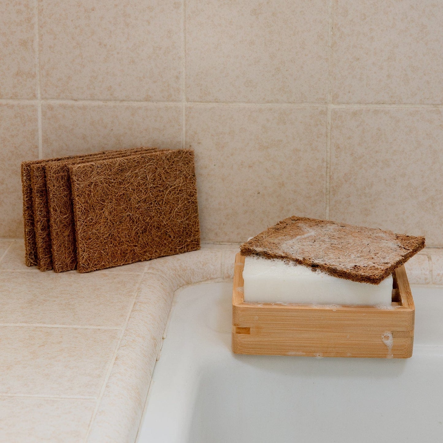 A bar of soap in a wooden dish, topped with a ZeroWasteStore.com Coconut Kitchen Scrubber, rests on the bathtub edge. Three more brown Coconut Kitchen Scrubbers are stacked on the beige tiled counter nearby.