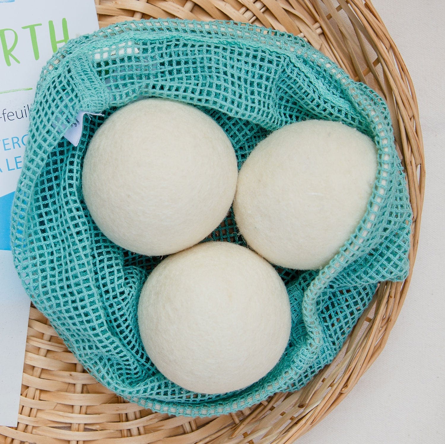 Three Reusable Wool Dryer Balls from ZeroWasteStore.com are in a light blue mesh bag on a woven basket, with part of their white and blue paper packaging visible, promoting their use as a natural fabric softener.