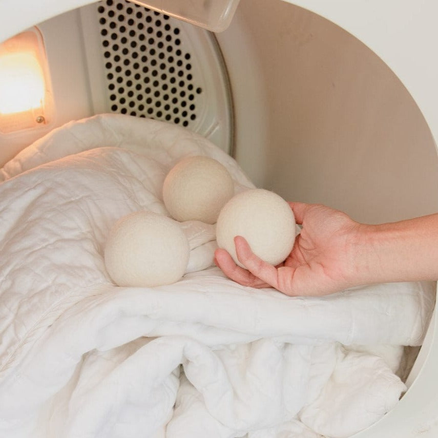 A hand places ZeroWasteStore.com Reusable Wool Dryer Balls on a white comforter inside a lit dryer, illuminating this natural fabric softener alternative.