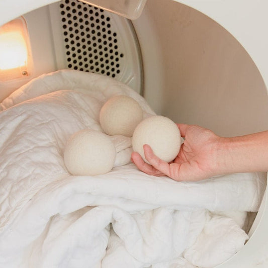 A hand places ZeroWasteStore.com Reusable Wool Dryer Balls on a white comforter inside a lit dryer, illuminating this natural fabric softener alternative.