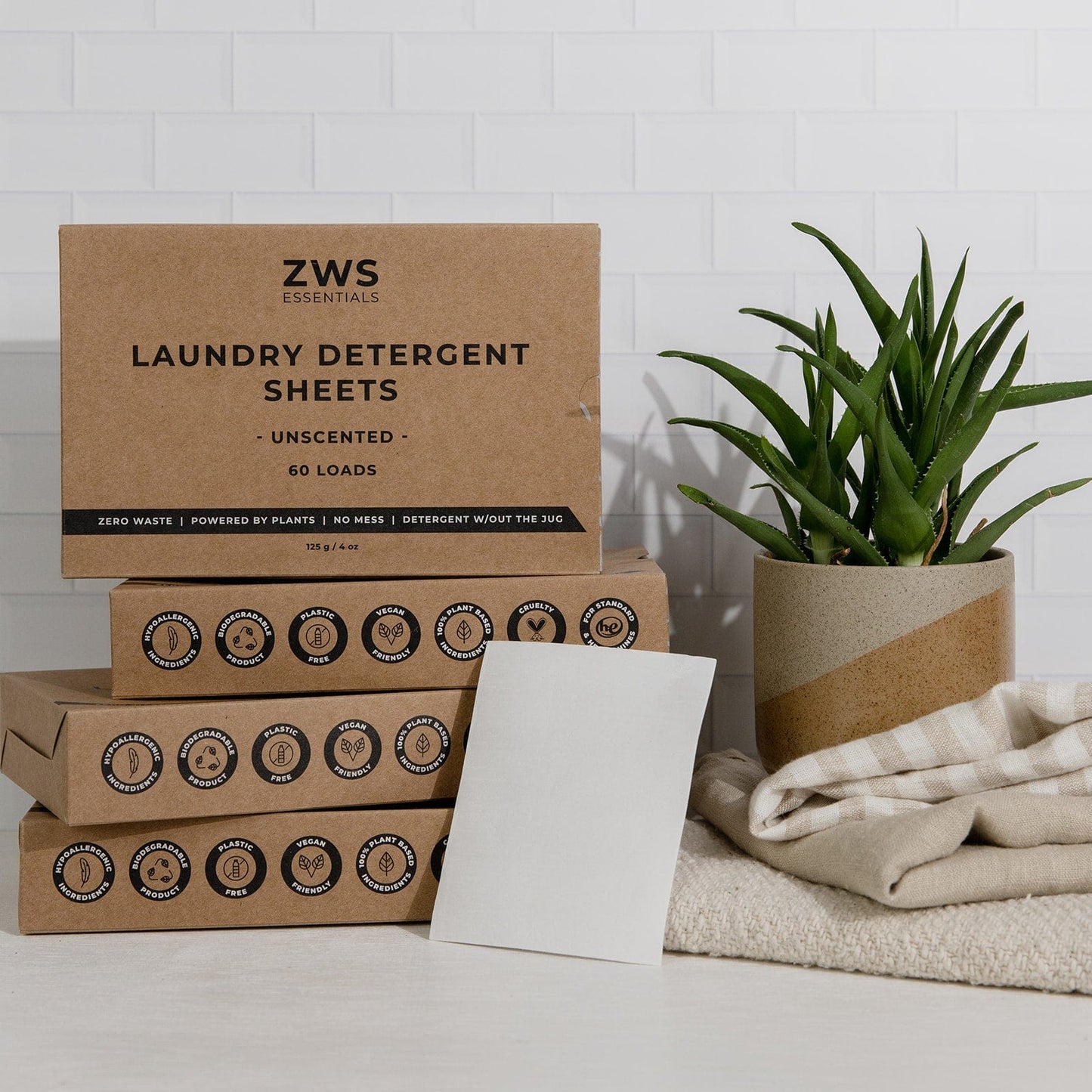 A stack of ZeroWasteStore.com Laundry Detergent Mini Kit boxes sits beside a potted plant, folded beige towel, and single white sheet, set against a white tiled background.