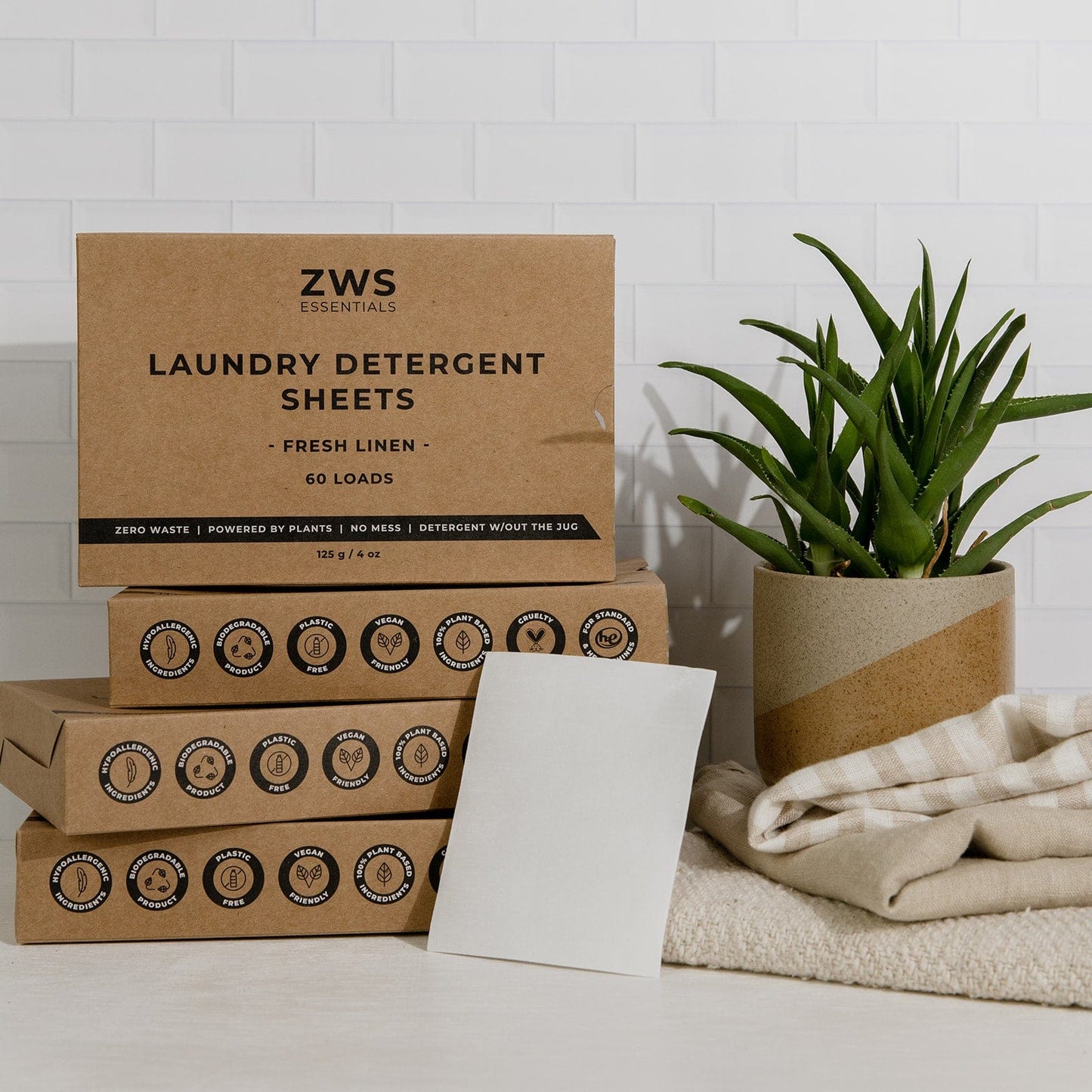 Four ZeroWasteStore.com Laundry Detergent Mini Kit boxes (2 or 4 pack) are stacked on a white surface next to a potted plant, folded towel, and a detergent sheet, with crisp white tiles in the background.