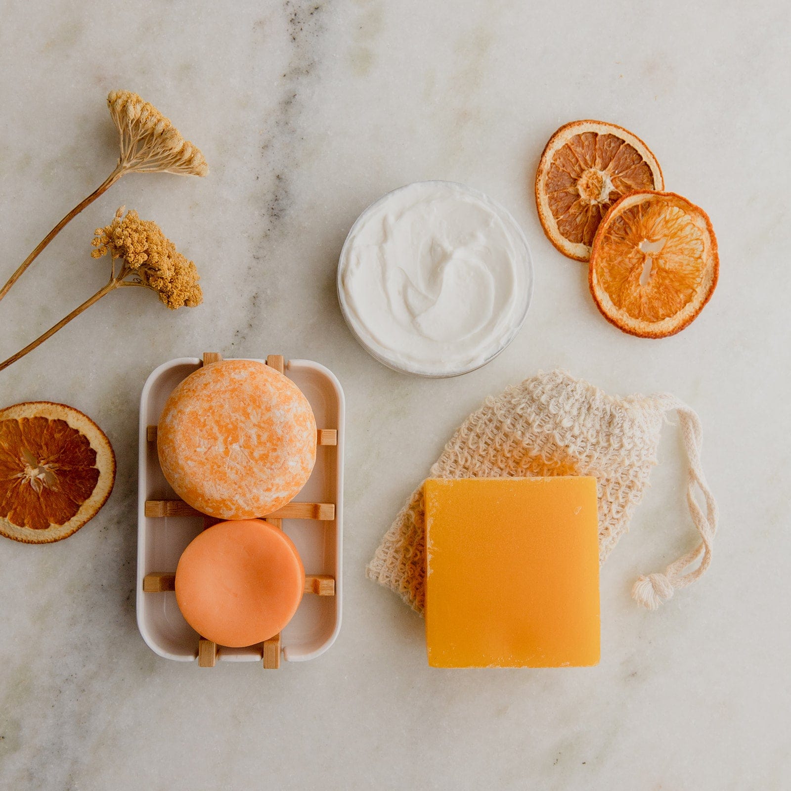 A flat lay of natural bath essentials from ZeroWasteStore.com, showcasing the Zero Waste Body Butter in Shampoo Bar Scents, dried orange slices and flowers, two round soaps on a tray, a yellow soap bar, white cream jar, and mesh soap pouch on marble.