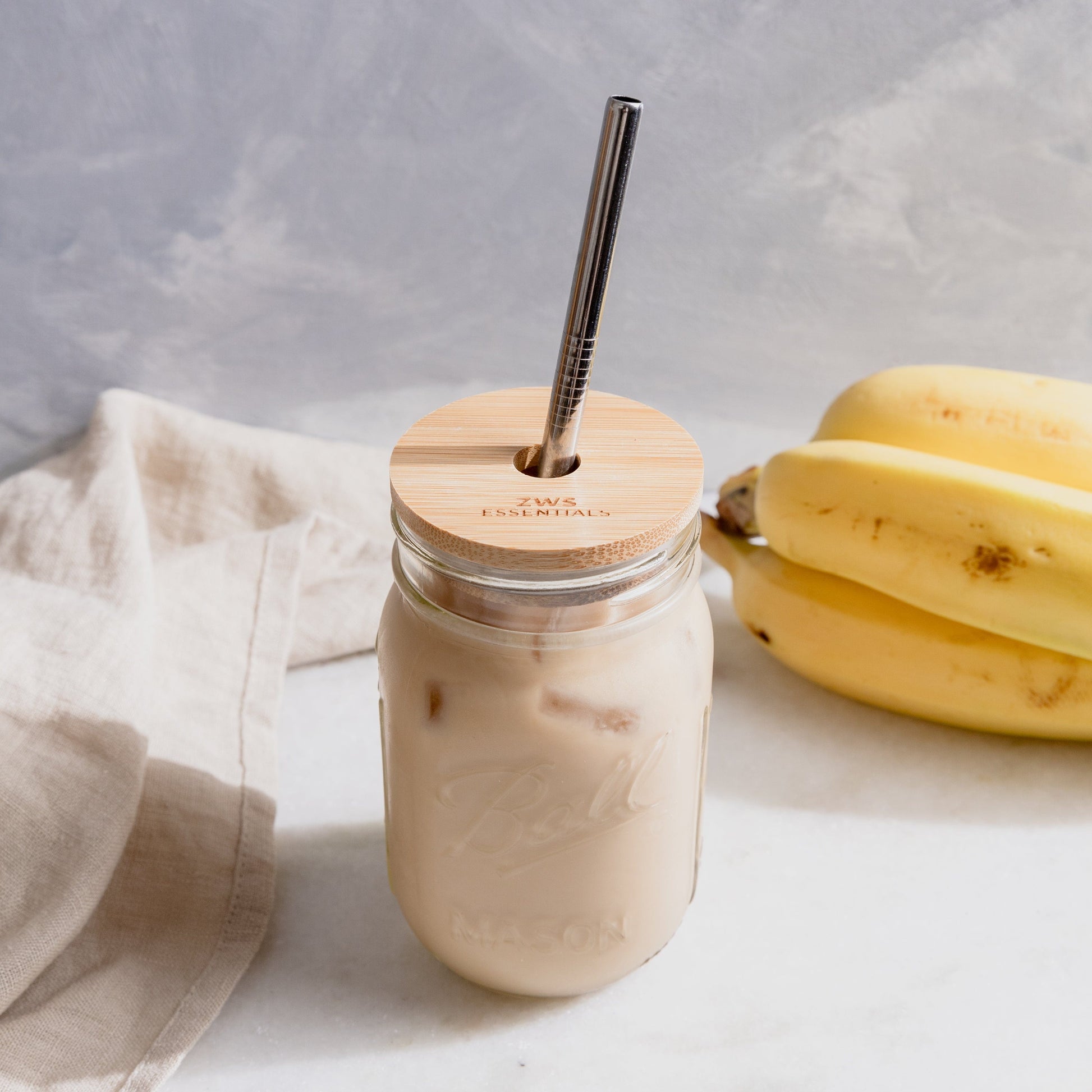 A Mason Jar Kit from ZeroWasteStore.com—with mason jar, bamboo lid, and stainless steel straw—sits on a white surface beside bananas and a beige cloth, filled with iced coffee or milk.