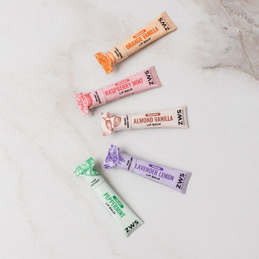 Five ZeroWasteStore.com All Natural Lip Balm tubes—Orange Vanilla, Raspberry Mint, Almond Vanilla, Lavender Lemon, and Peppermint—are arranged diagonally on a light marble surface.