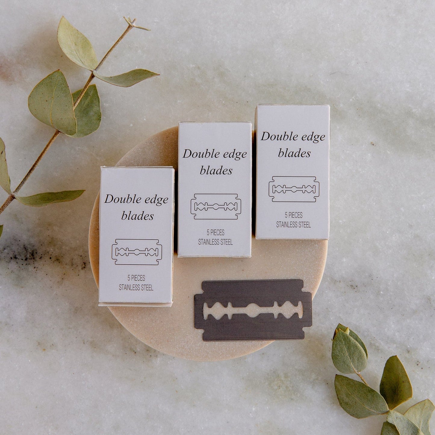Three boxes of ZeroWasteStore.com’s Zero Waste Razor Replacement Blades and a single blade rest on a dish, with green eucalyptus leaves on marble—an eco-friendly addition to your sustainable shaving routine.