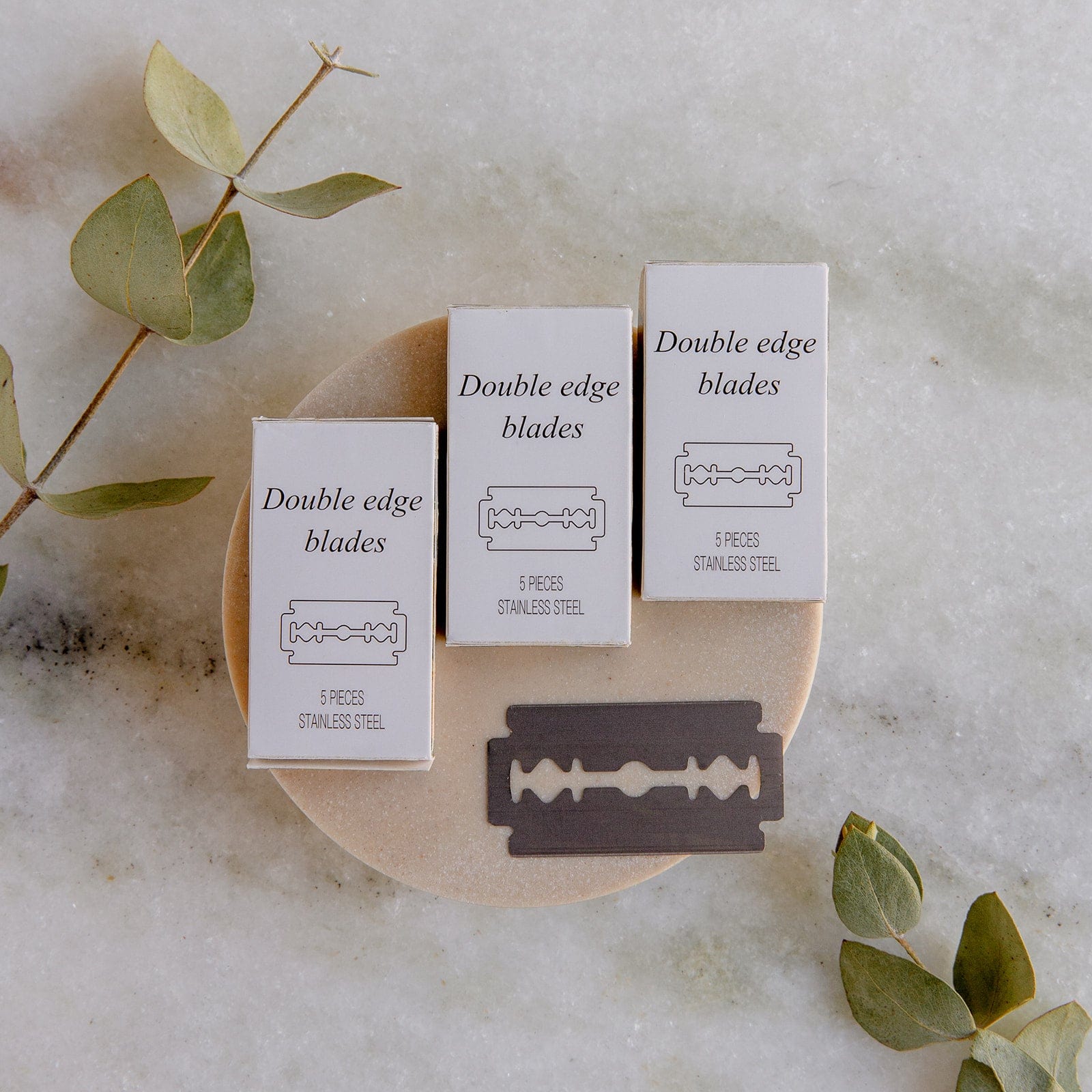 Three boxes of ZeroWasteStore.com’s Zero Waste Razor Replacement Blades and a single blade rest on a dish, with green eucalyptus leaves on marble—an eco-friendly addition to your sustainable shaving routine.