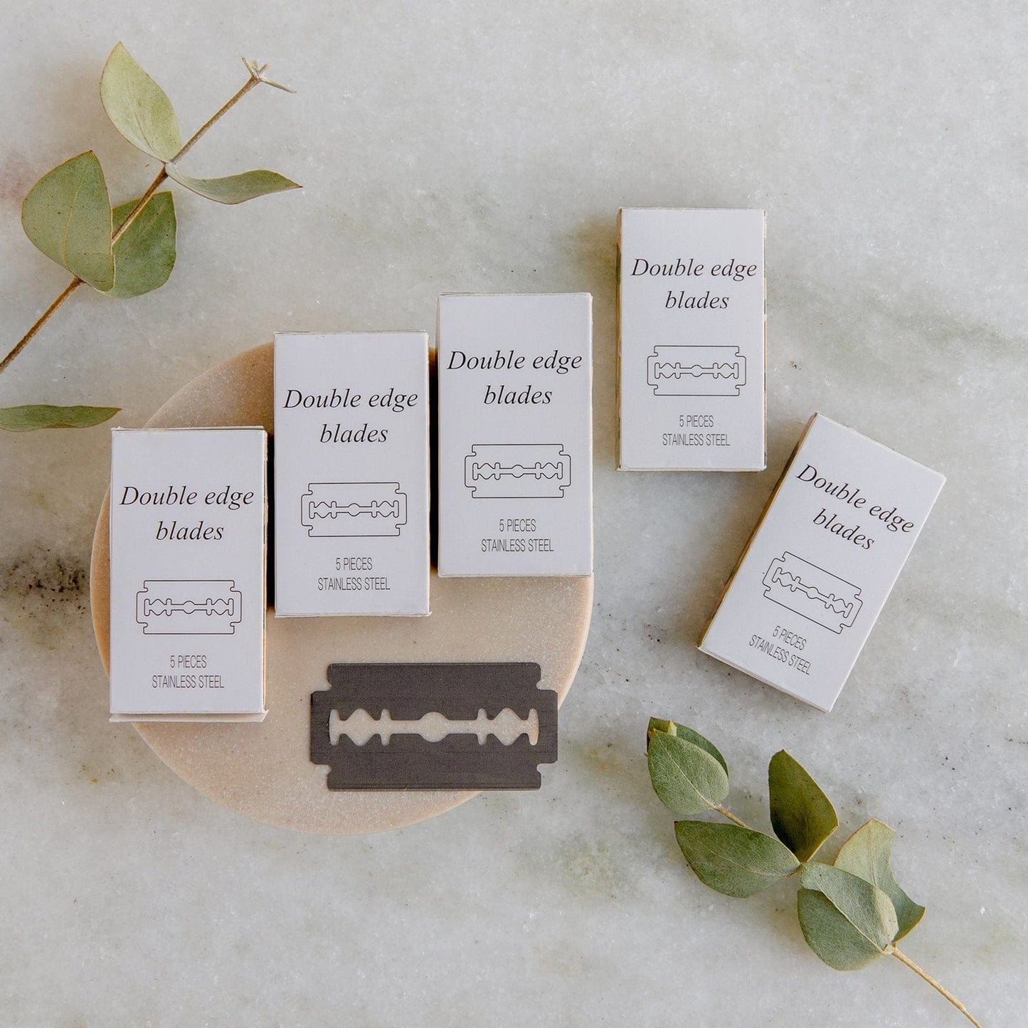 Five boxes of ZeroWasteStore.com Zero Waste Razor Replacement Blades and a single blade rest on a white round surface with eucalyptus leaves scattered nearby—a stylish zero-waste shaving setup on marble.