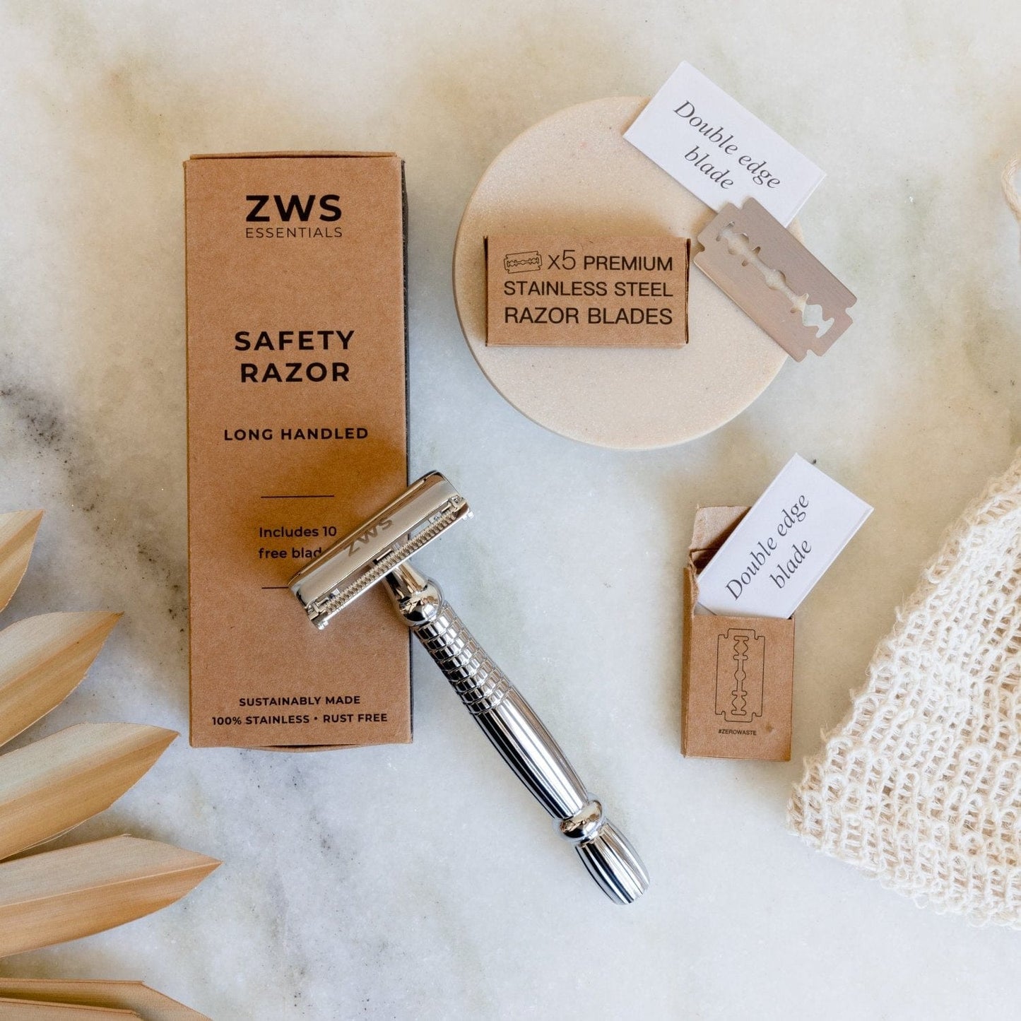 A ZeroWasteStore.com Zero Waste Razor, Replacement Blades, a cardboard razor box, beige soap dish, and mesh bag are displayed on a white surface next to a wooden plant decoration.