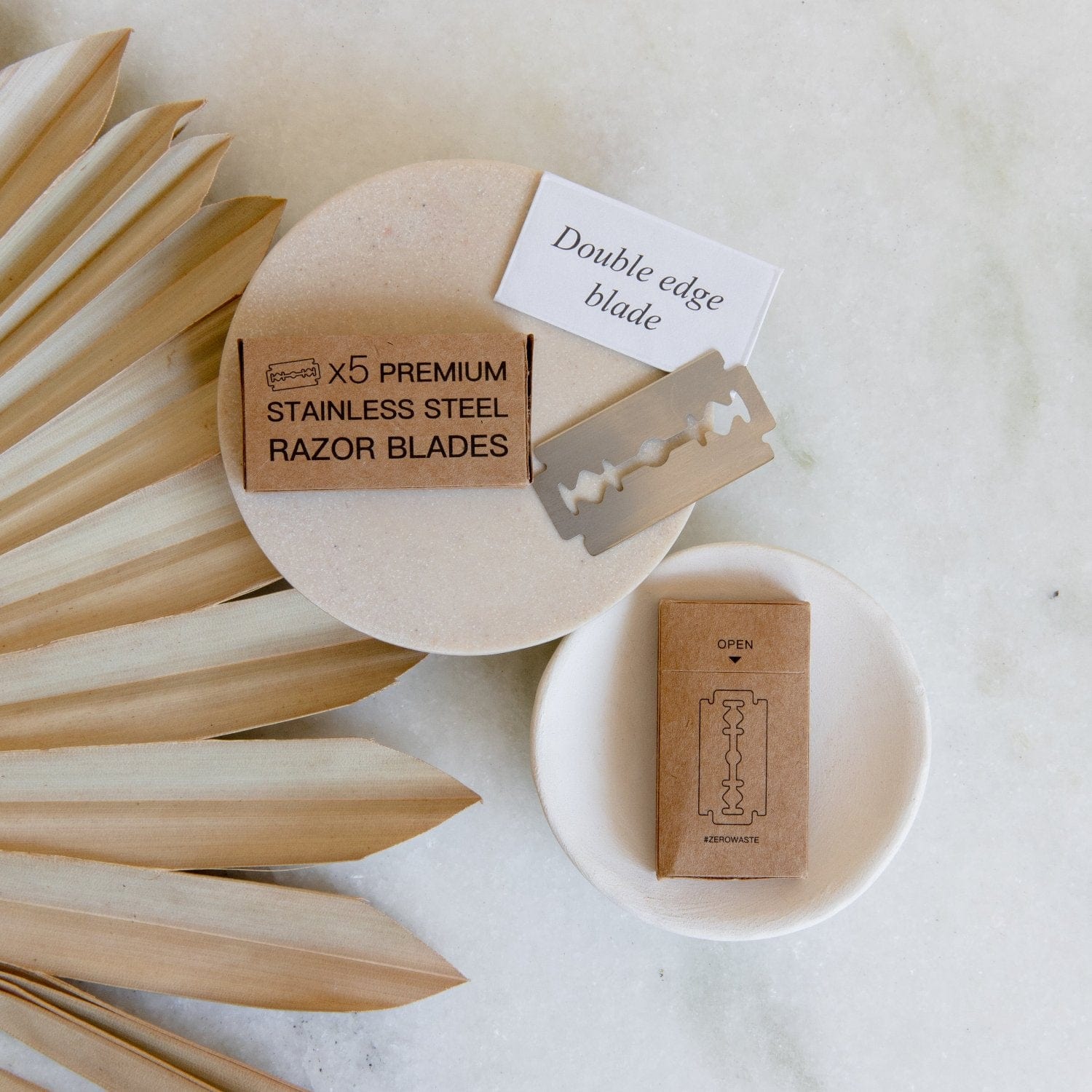 A flat lay features Zero Waste Razor Replacement Blades by ZeroWasteStore.com in cardboard packaging, an individual stainless steel blade, and dried palm leaves on marble—minimal, eco-friendly essentials for zero waste shaving routines.