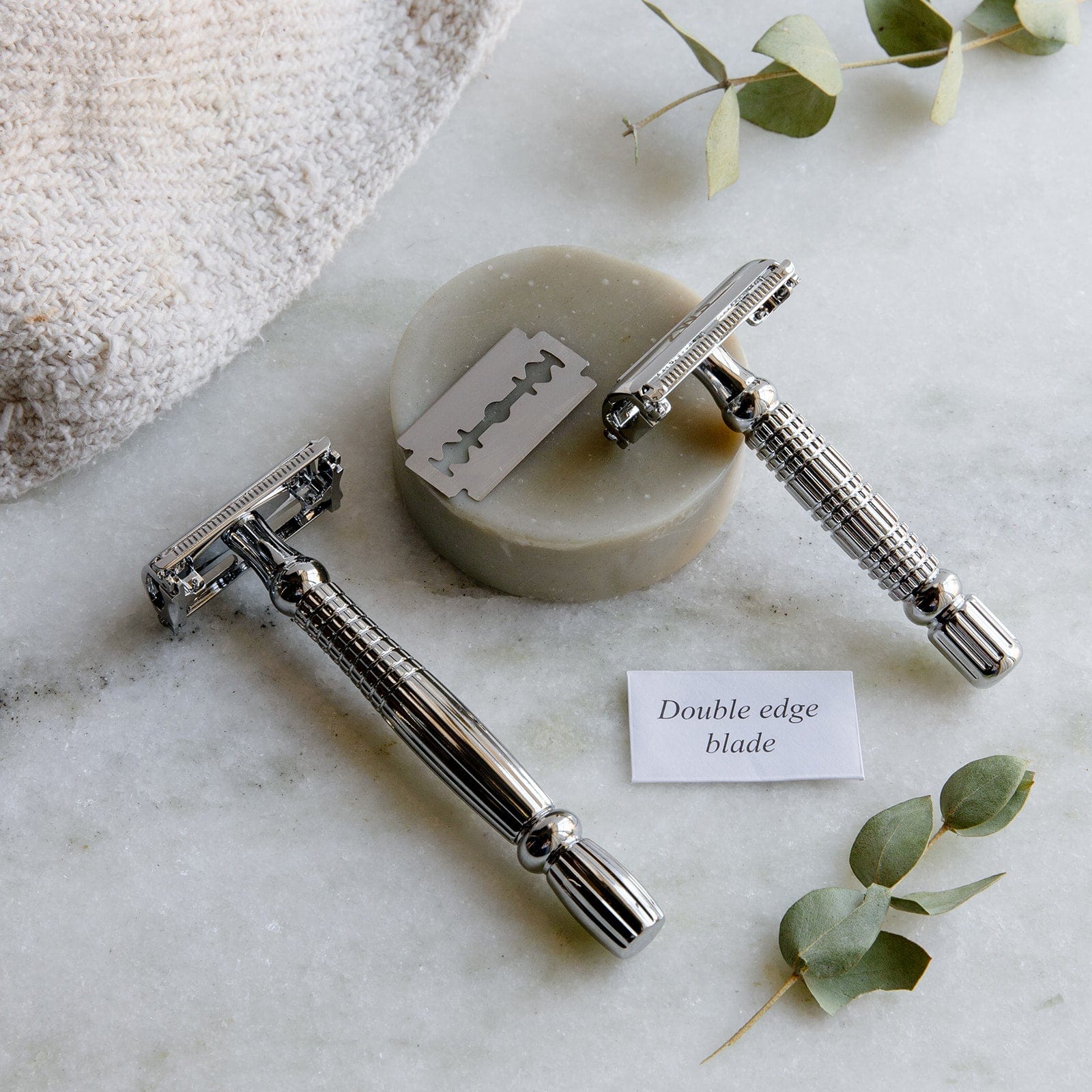 Two Zero Waste Safety Razors by ZeroWasteStore.com with 10 free blades, a round soap bar, towel, and green leaves rest on marble. A label highlights: “Double edge blade—ideal for zero plastic waste grooming.”.