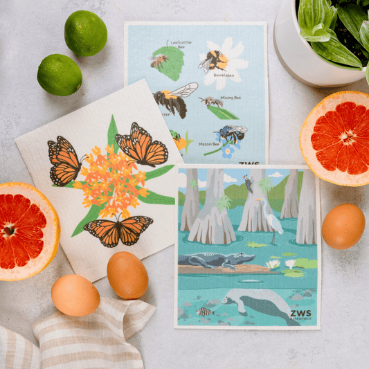 Two ZeroWasteStore.com Swedish Dish Cloths with butterfly, bee, and wetland animal designs rest on a countertop with grapefruit, limes, eggs, and a potted plant—a colorful, eco-friendly paper towel alternative.