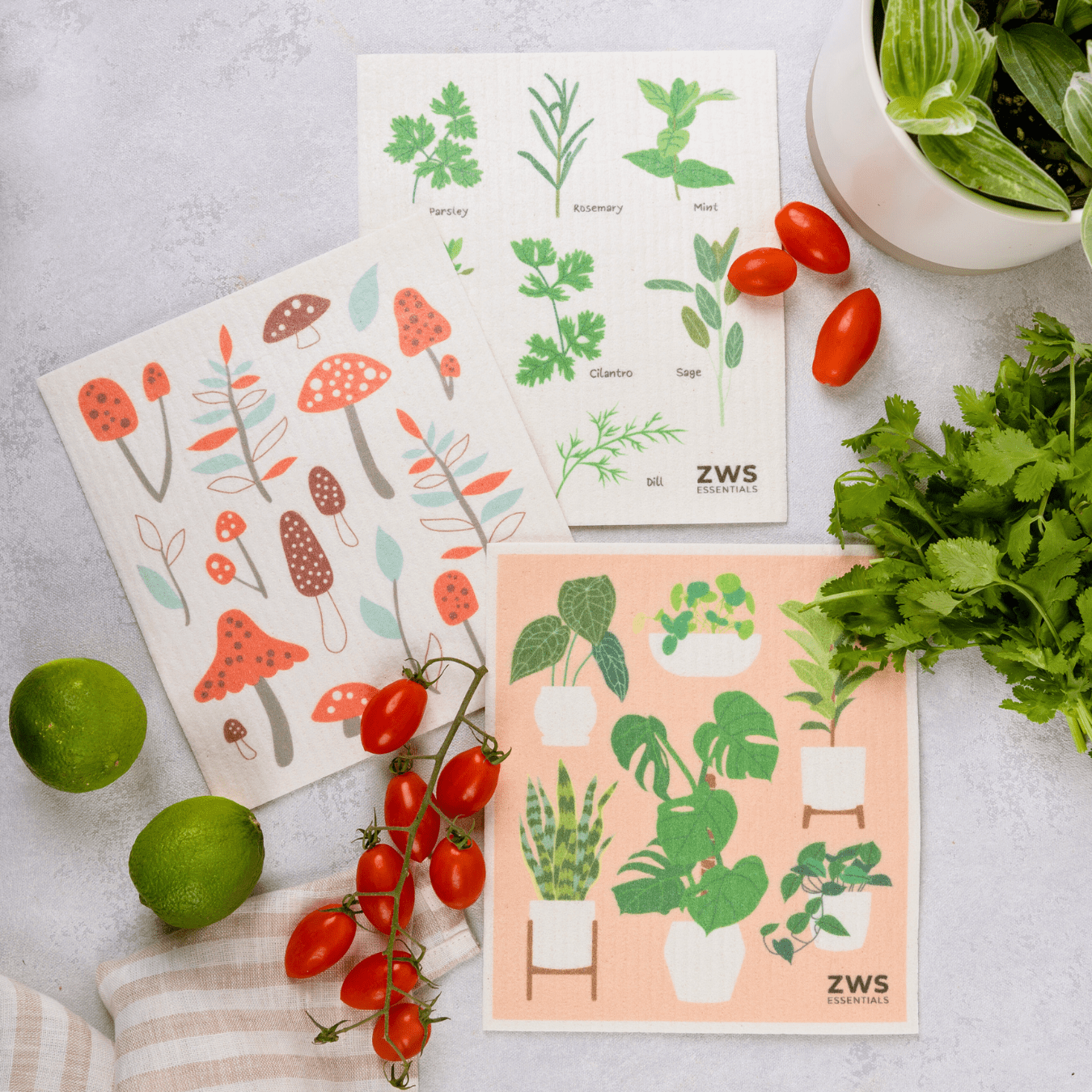 Three ZeroWasteStore.com Swedish Dish Cloths with plant and mushroom designs are displayed on a light surface amid cherry tomatoes, limes, cilantro, and a napkin—an eco-friendly paper towel replacement.