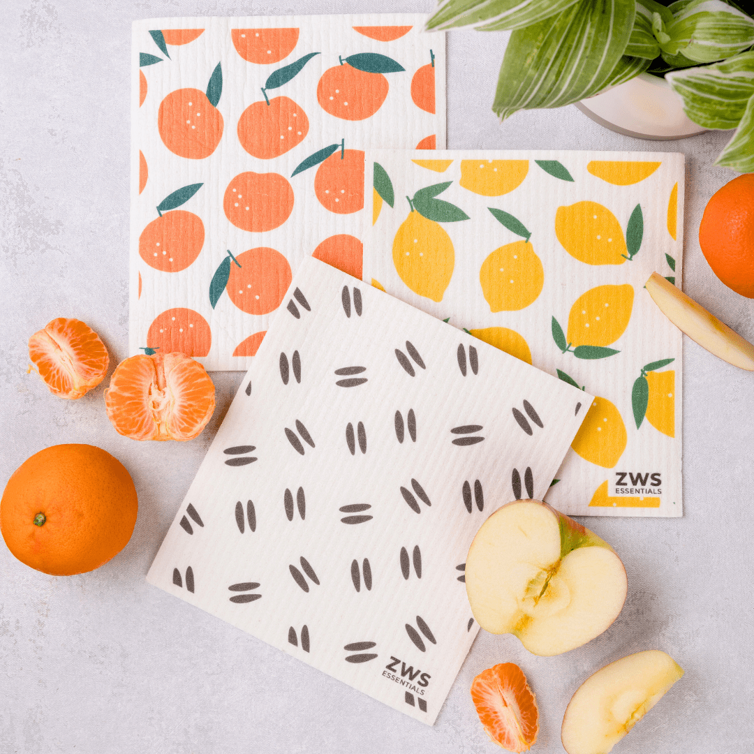 Three Swedish Dish Cloths from ZeroWasteStore.com, featuring orange, lemon, and seed patterns, are displayed with fruit and a plant—an eco-friendly paper towel alternative for zero waste cleaning.