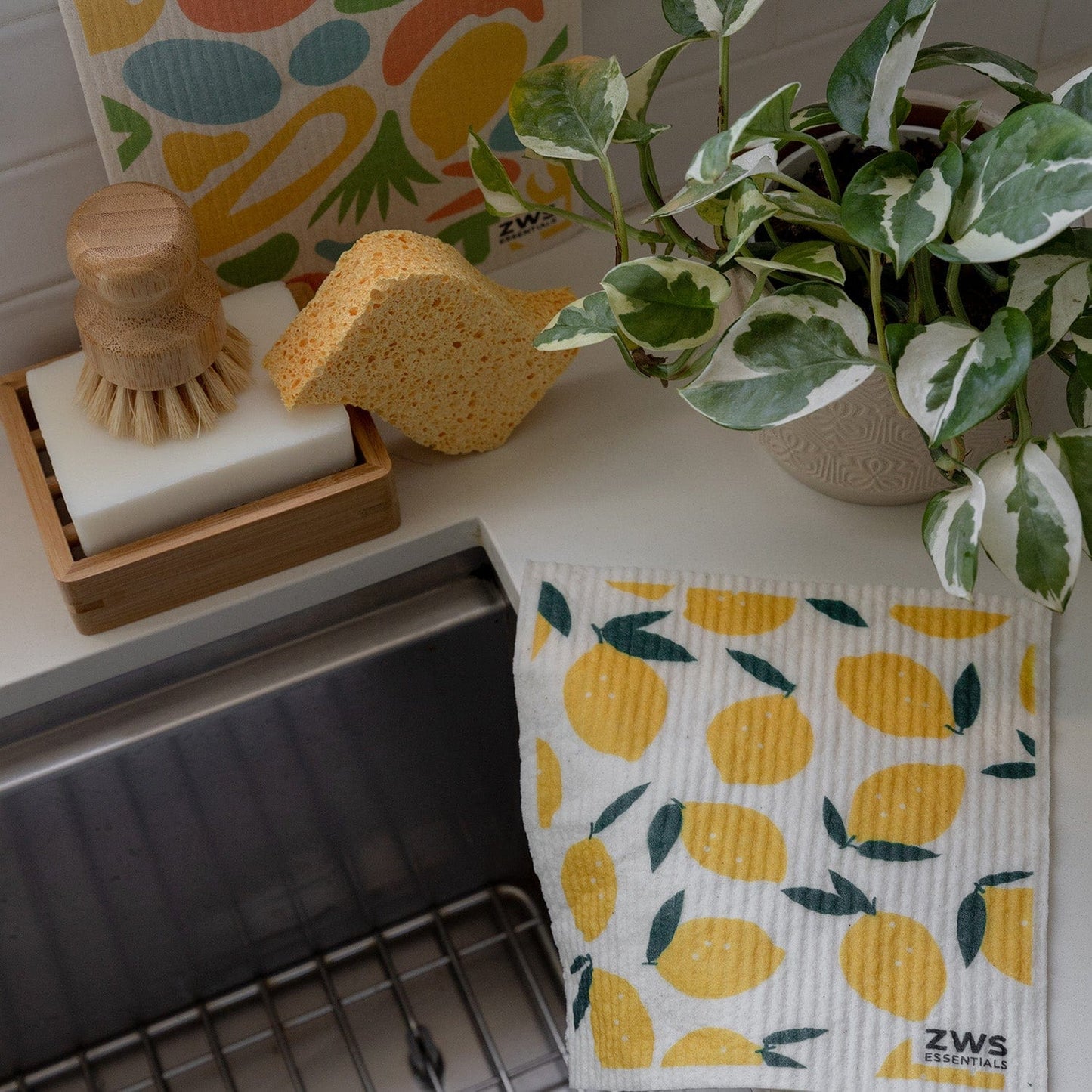A kitchen sink area with a potted variegated plant, a scrub brush in a soap dish, and colorful dishcloths—including the Swedish Dish Cloth from ZeroWasteStore.com with lemons and an abstract fruit design sponge cloth.