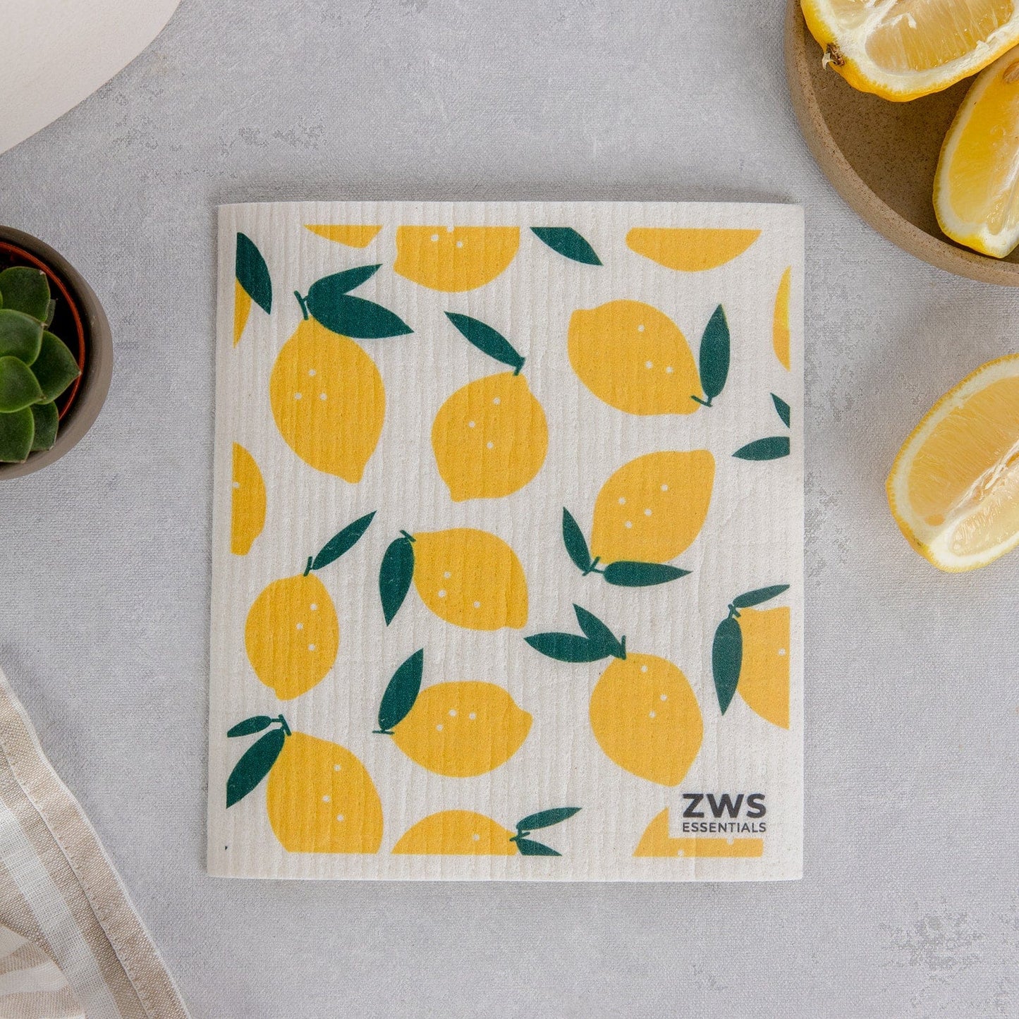 A reusable Swedish Dish Cloth from ZeroWasteStore.com, featuring a yellow lemon and green leaf pattern, rests on a light gray surface with lemon slices, a small plant, and a plaid cloth—an eco-friendly paper towel alternative.