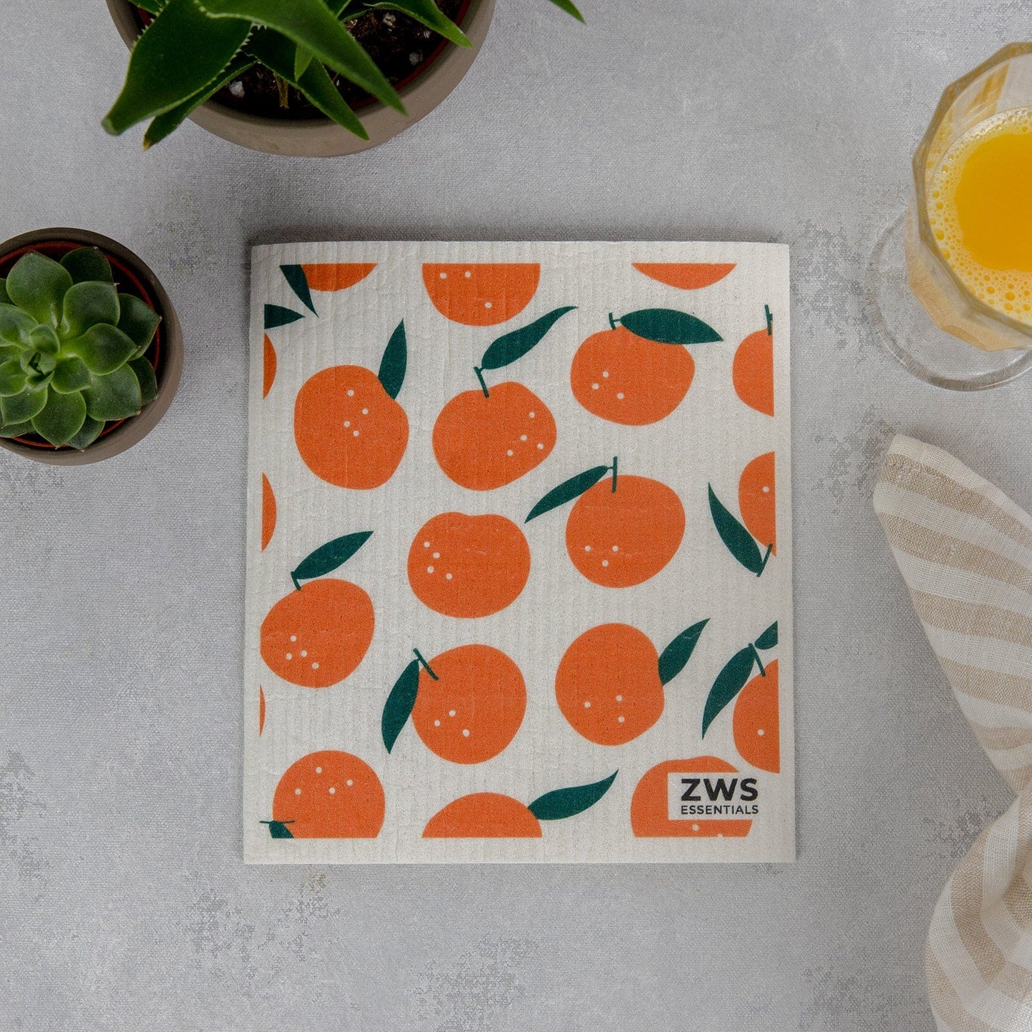 A Swedish Dish Cloth from ZeroWasteStore.com, featuring orange fruit and green leaf patterns, rests on a gray surface beside a succulent, orange juice glass, striped napkin, and has the brand marked in one corner.