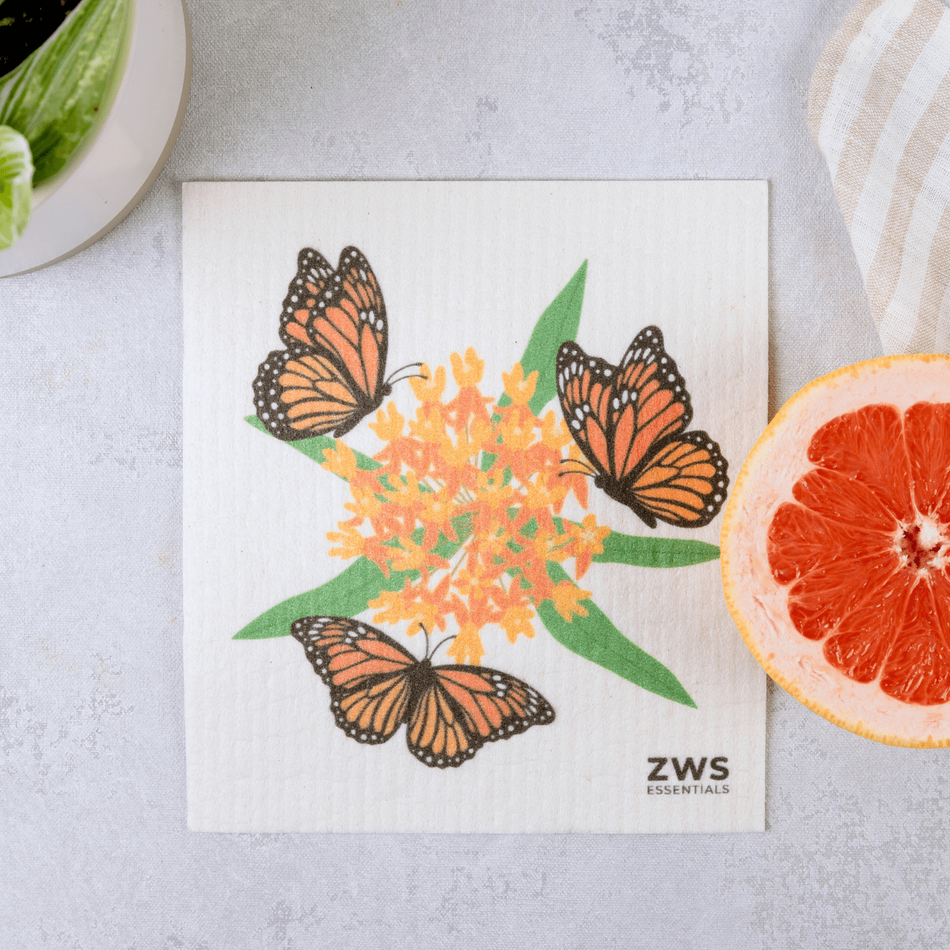 A square Swedish Dish Cloth from ZeroWasteStore.com, featuring three monarch butterflies around orange flowers, sits by a grapefruit half, potted plant, and striped cloth—the perfect eco-friendly paper towel replacement.