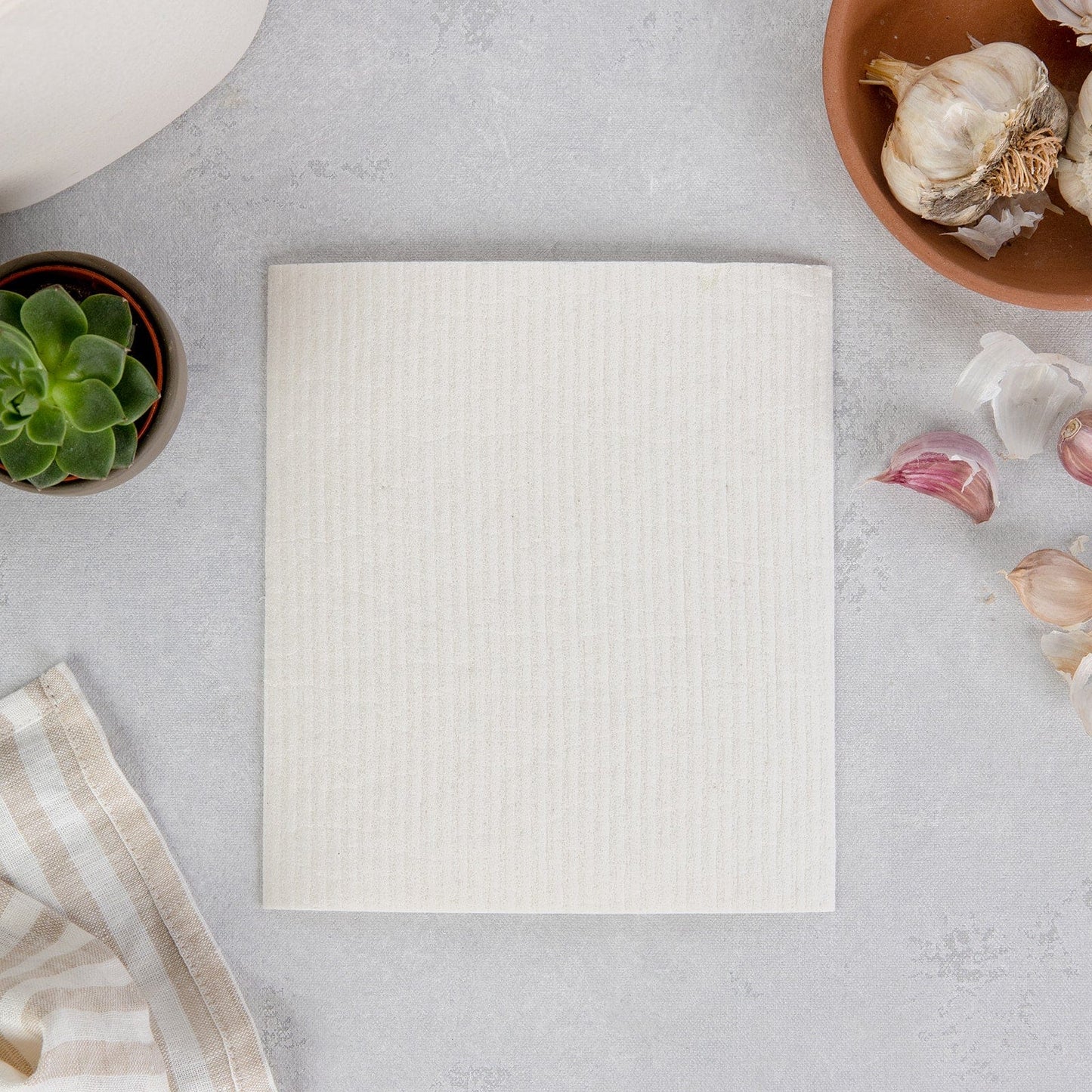 A beige folded napkin sits on gray beside a succulent, striped cloth, garlic, and cloves—showcasing the Swedish Dish Cloth by ZeroWasteStore.com as an eco-friendly, stylish paper towel alternative.