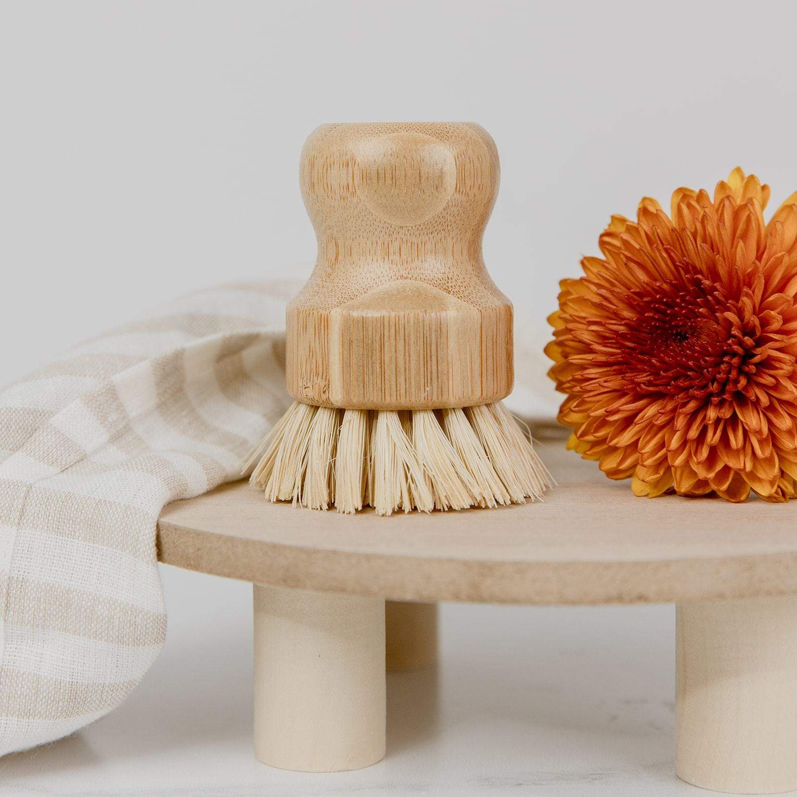 The ZeroWasteStore.com Dish Brush with natural bristles stands on a round wooden platform beside an orange chrysanthemum and a folded beige-striped cloth, creating a chic, plastic-free cleaning setup.