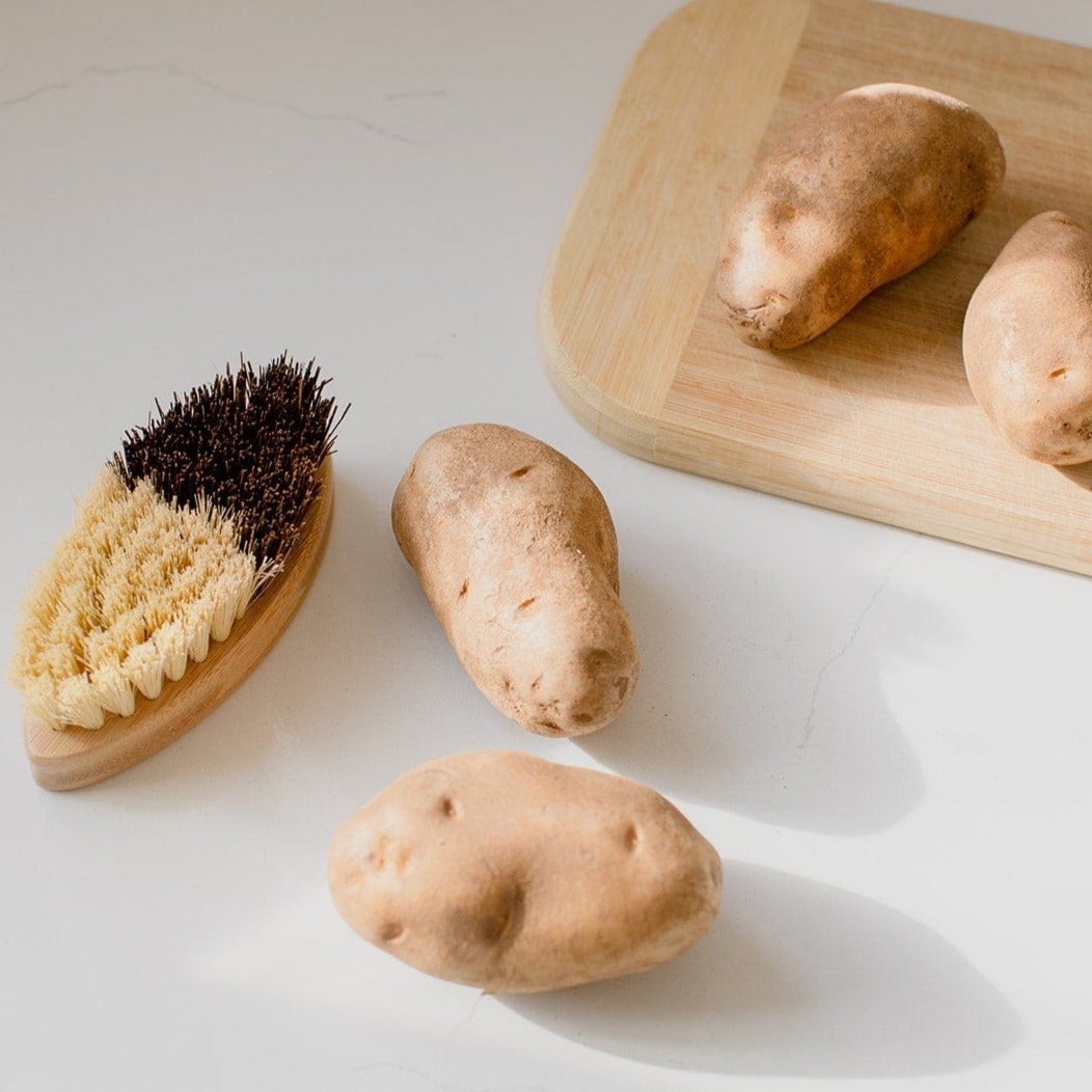 A small ZeroWasteStore.com Vegetable Brush and three raw potatoes are on a white surface, with another potato on a wooden cutting board in the background.