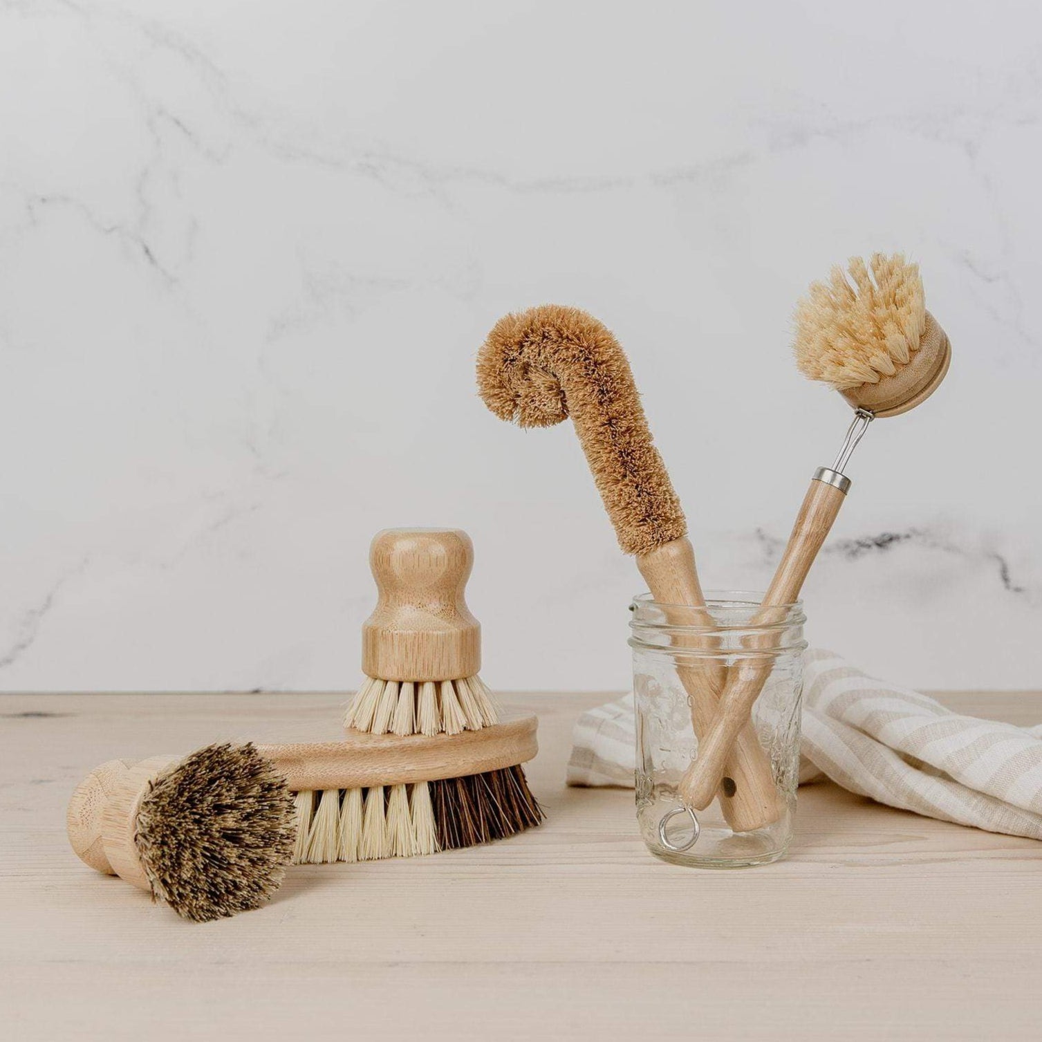 The Zero Waste Dish Brush Kit from ZeroWasteStore.com features four wooden biodegradable brushes with natural bristles, arranged on a light wood surface—two in a glass jar and a beige striped cloth nearby against a light background.