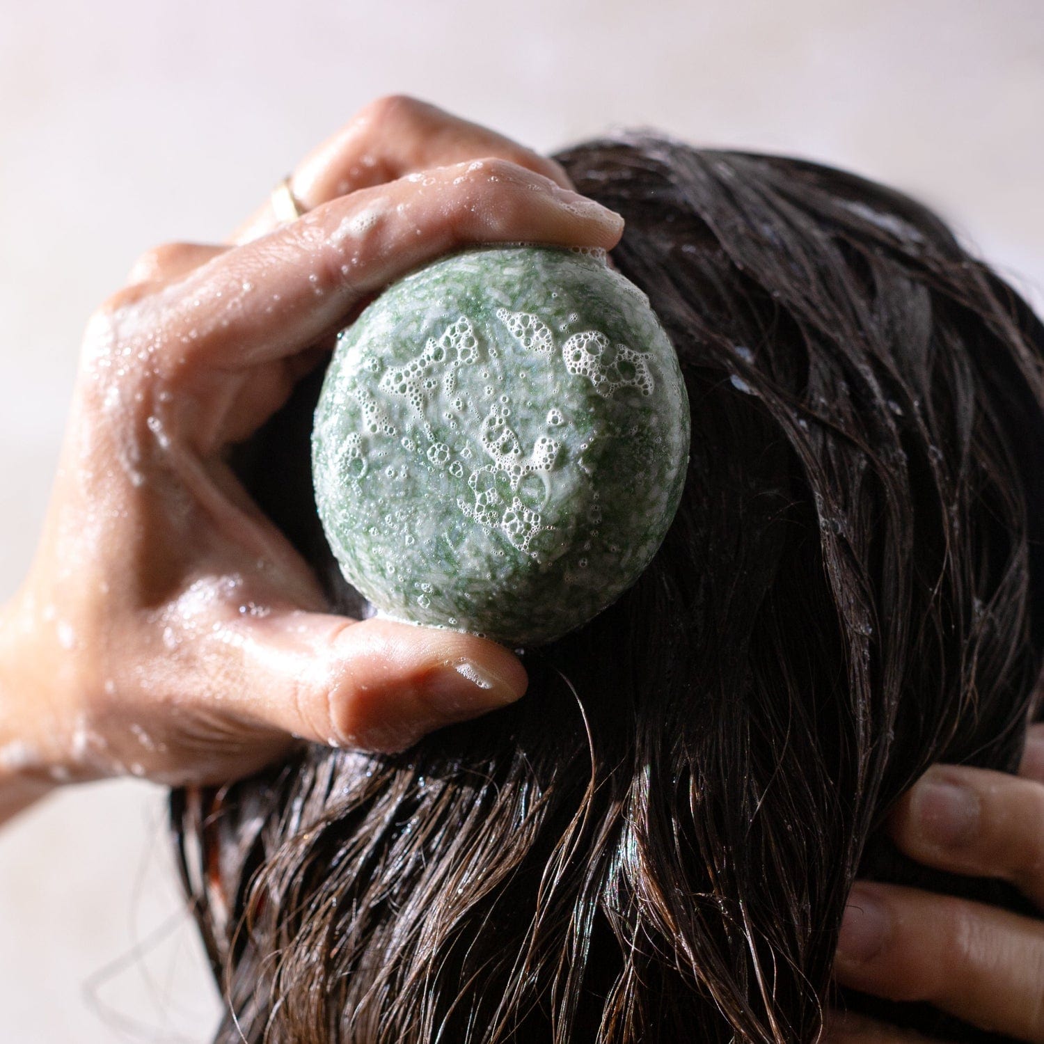 A hand rubs the green, bubbly Shampoo Bar from ZeroWasteStore.com onto wet, dark hair, creating a rich vegan lather and foam.