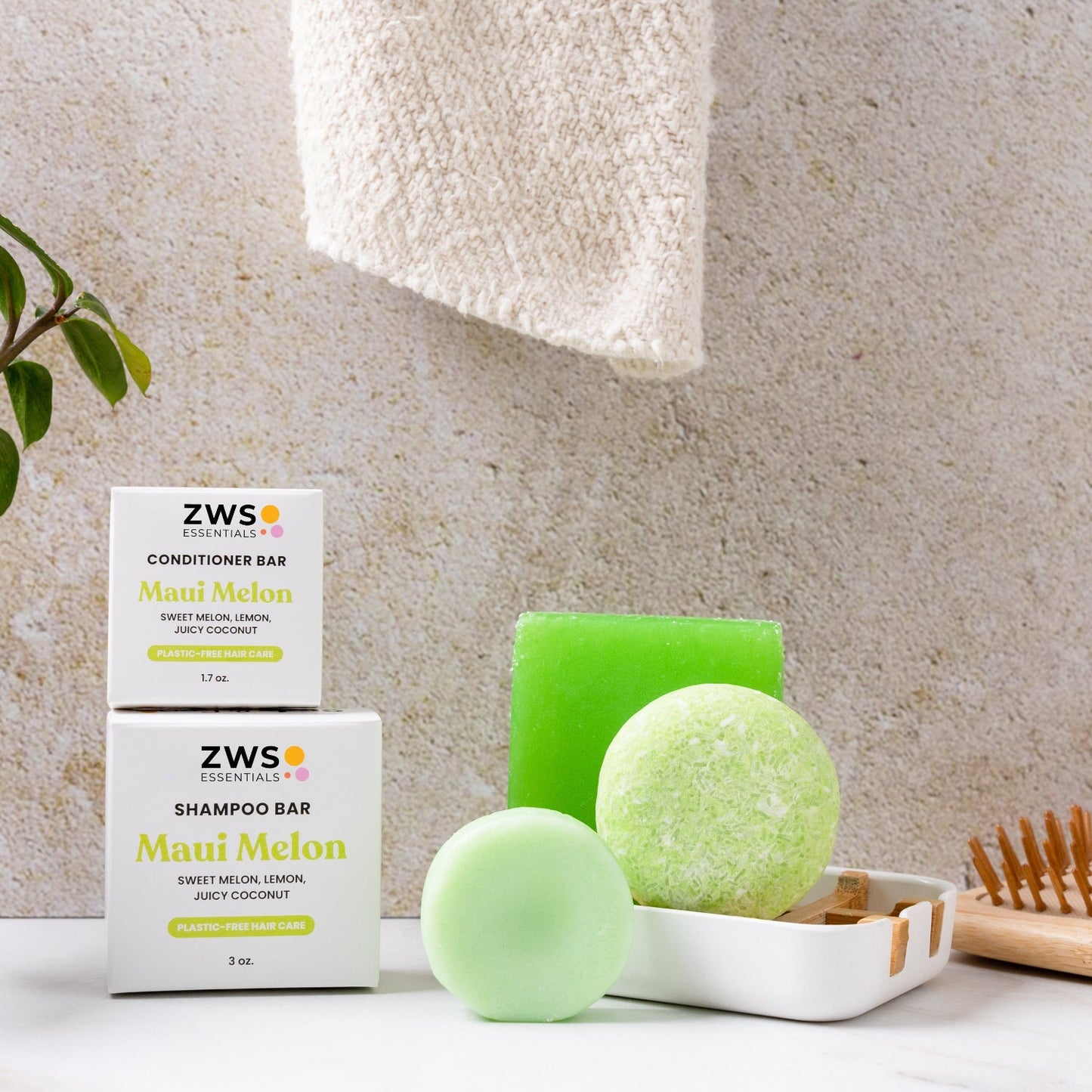 A green Shampoo Bar from ZeroWasteStore.com is displayed with its Maui Melon box, alongside a conditioner bar and soap. A towel hangs above, with a plant and toothbrush nearby—ideal for an eco-friendly haircare routine.