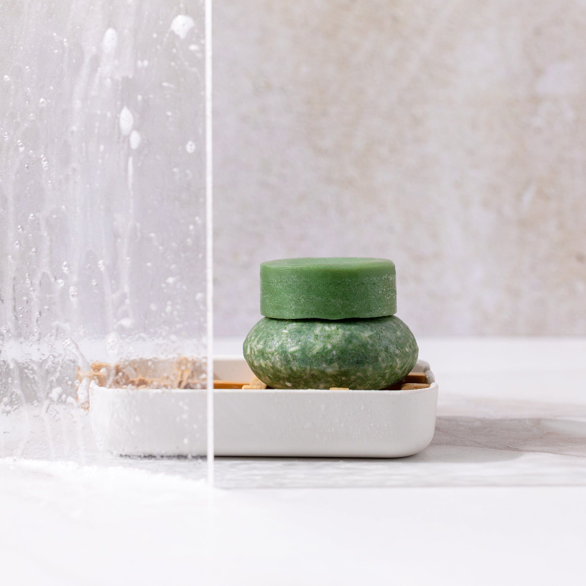 Bloom Shampoo & Conditioner Bar Duo by ZeroWasteStore.com, made with castor oil for hair growth, sits stacked on a white soap dish with water splashes nearby. The solid bars stand out against a light, neutral background.