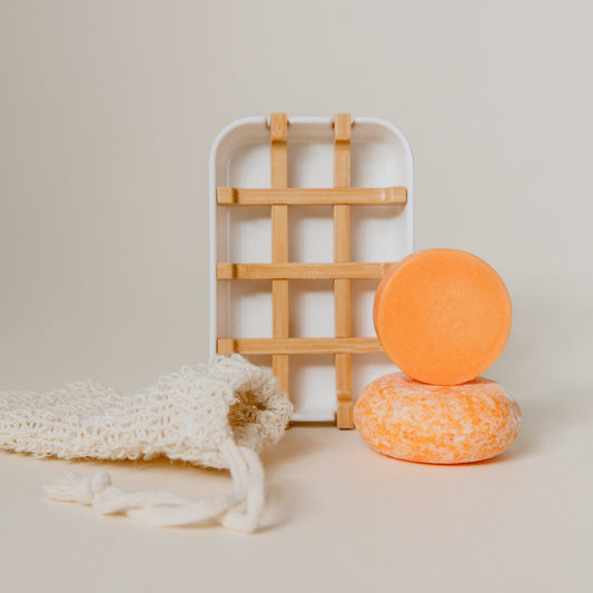 The Zero Waste Hair Care Mini Kit from ZeroWasteStore.com features two round orange shampoo bars, a white and bamboo soap dish, and an off-white mesh pouch—perfect for a sustainable bathroom routine.