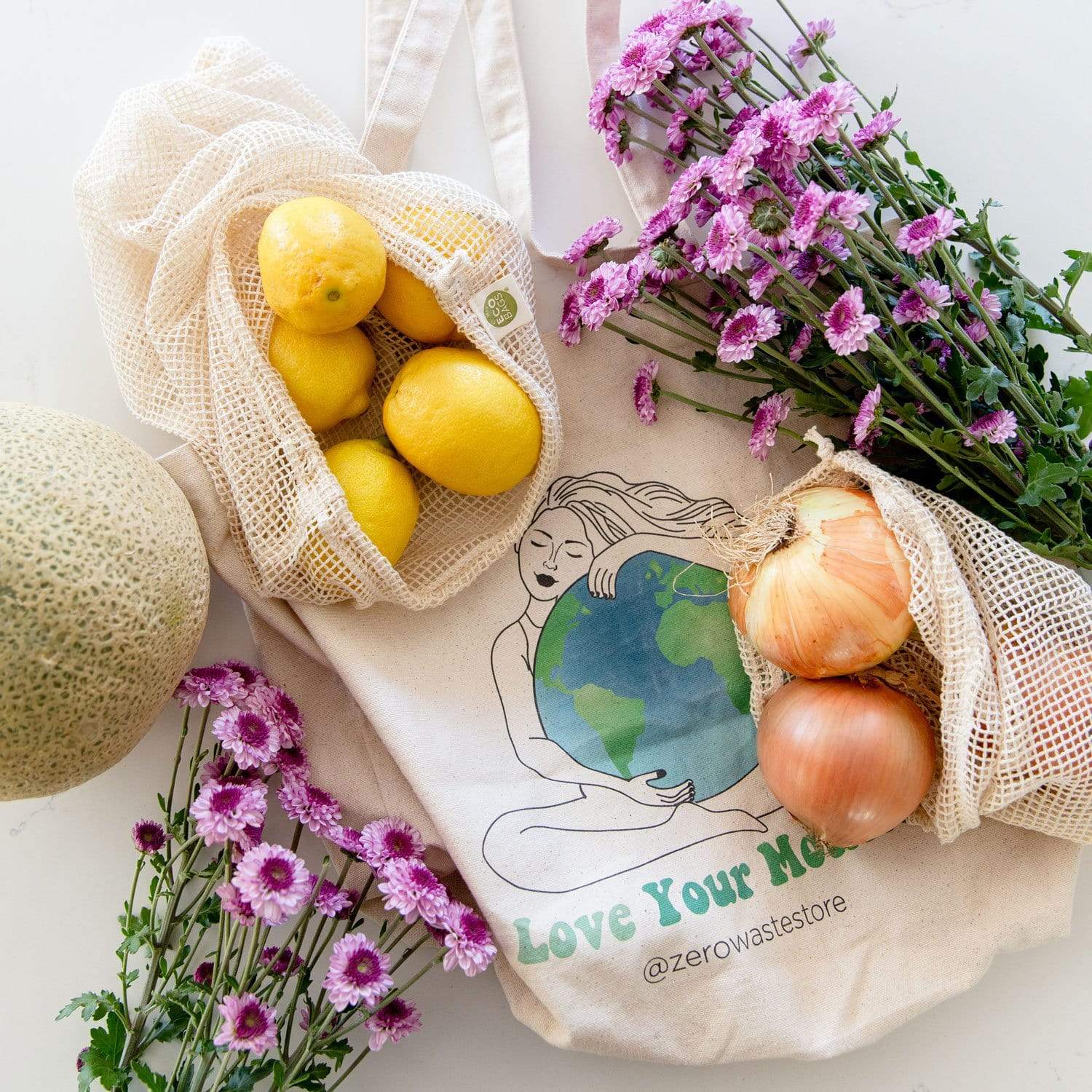 The Zero Waste Tote Bag Kit by ZeroWasteStore.com includes reusable produce bags with yellow lemons, purple flowers, and onions on a white surface. One organic cotton bag shows a woman hugging Earth with “Love Your Mother @zerowastestore.”.