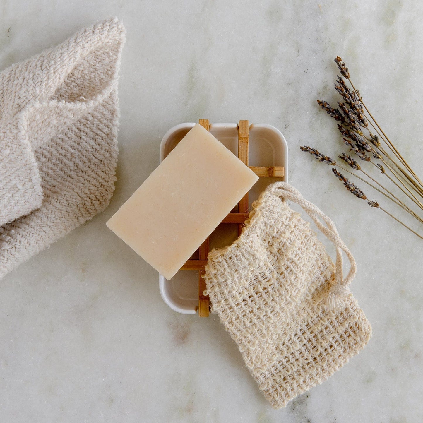 The Soap Bar Mini Kit from ZeroWasteStore.com, featuring a beige vegan bar soap, a textured washcloth, mesh soap saver pouch, and dried lavender, is displayed on a light marble surface.