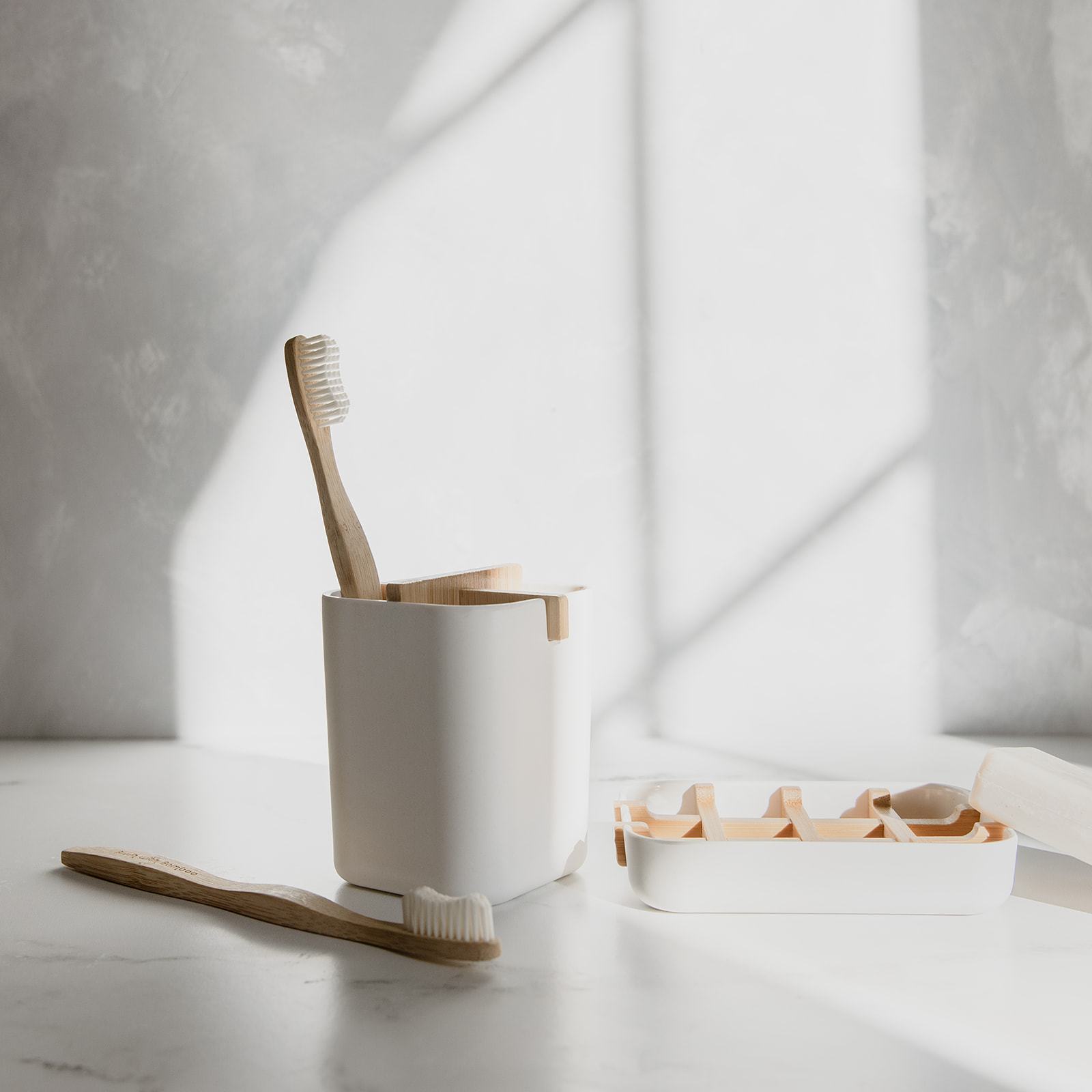 Two ZeroWasteStore.com Bamboo Toothbrushes—one upright in a white holder, one lying on a white surface—are shown with a separate white tray. Sunlight casts soft shadows across the minimalist scene.