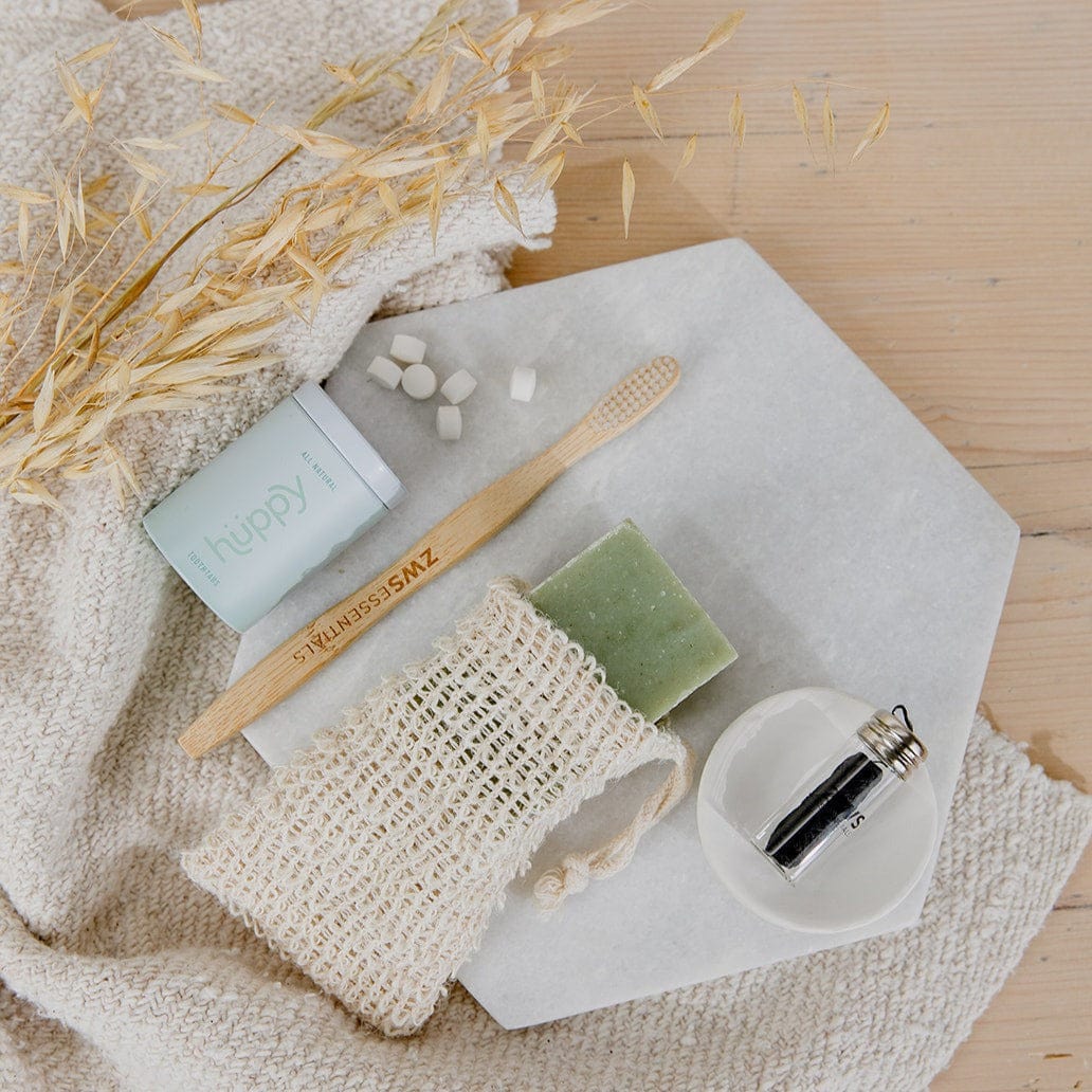 ZeroWasteStore.com's Bamboo Toothbrush, toothpaste tablets, a green soap bar in a mesh bag, a glass jar with black contents, and dried oat stems are displayed as eco-friendly bathroom essentials on cream cloth atop a white stone surface.