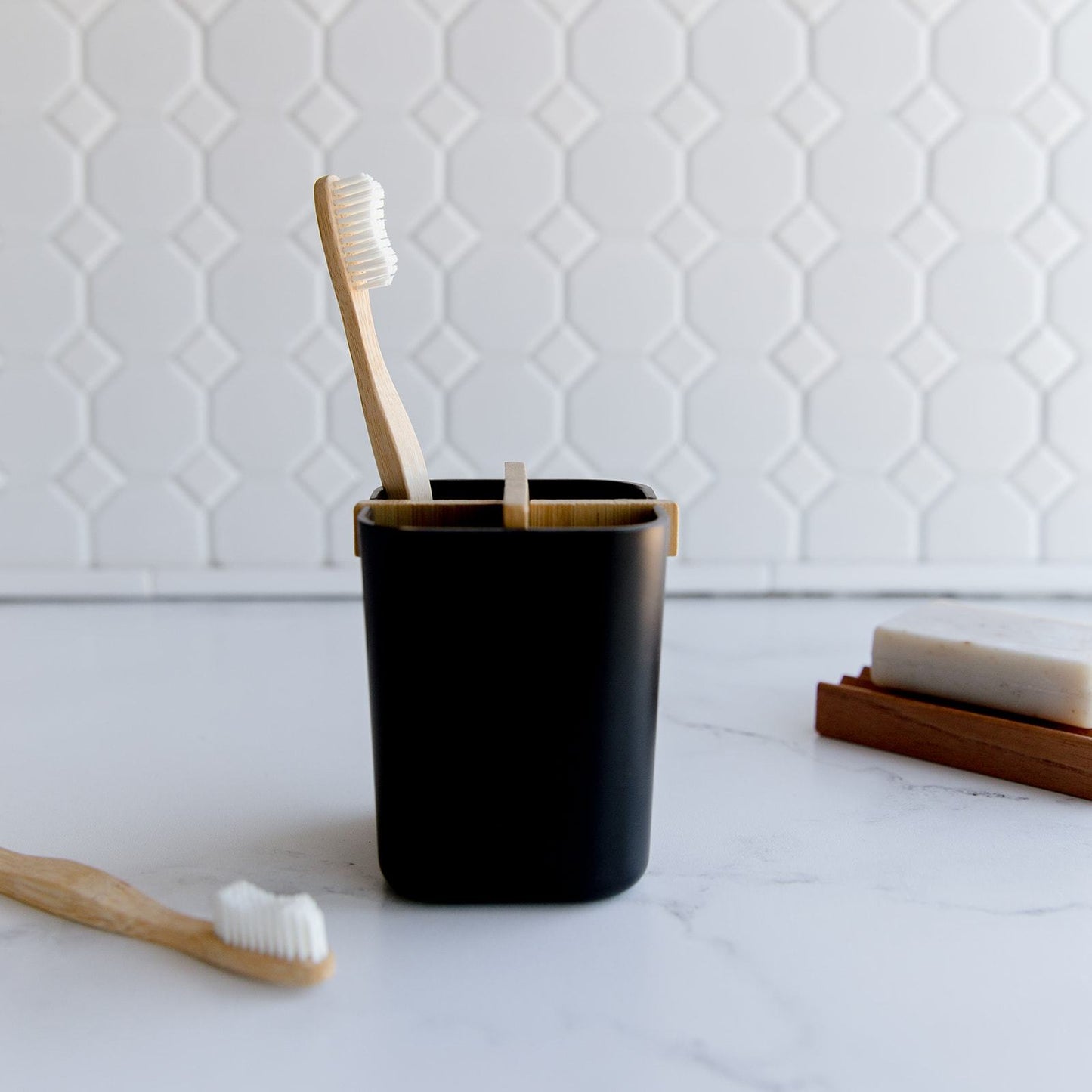 Two ZeroWasteStore.com Bamboo Toothbrushes in a black holder rest on a white marble counter, paired with a bar of soap on a wooden tray, all set against geometric tiles for a plastic-free routine.