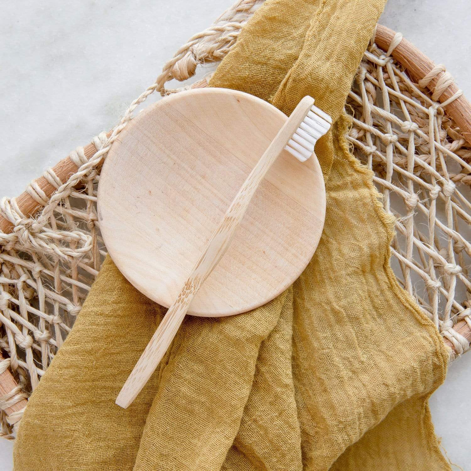 A Bamboo Toothbrush - Child by ZeroWasteStore.com rests on a round wooden soap dish atop a woven basket, with a mustard yellow cloth draped beneath.