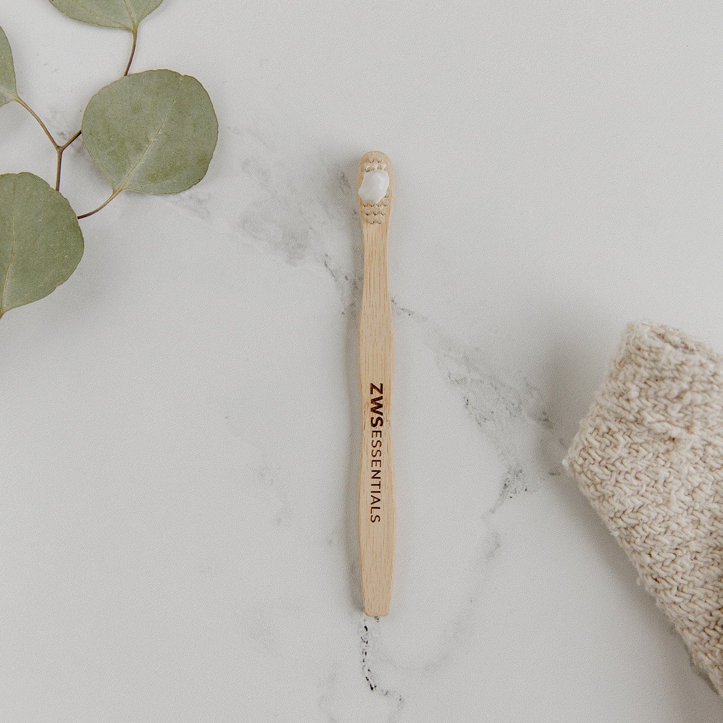 A Bamboo Toothbrush - Child from ZeroWasteStore.com with white bristles rests on a white marble surface next to green leaves and a knitted beige cloth.
