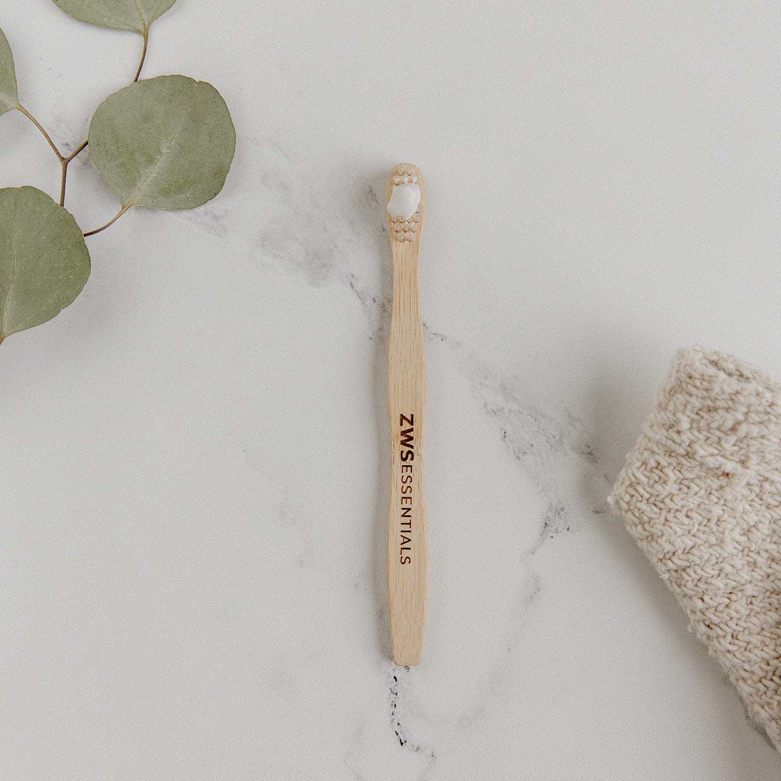 A Bamboo Toothbrush - Child from ZeroWasteStore.com with white bristles rests on a white marble surface next to green leaves and a knitted beige cloth.