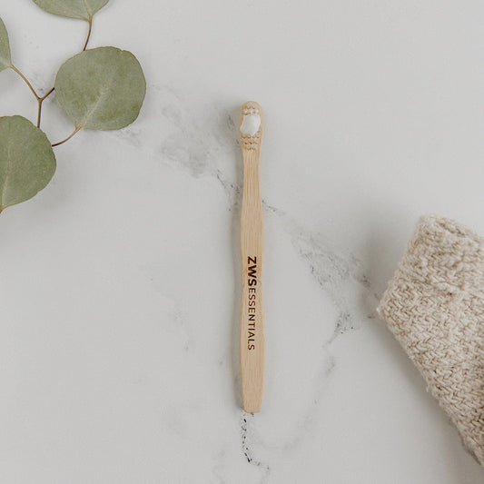 A Bamboo Toothbrush - Child from ZeroWasteStore.com with white bristles rests on a white marble surface next to green leaves and a knitted beige cloth.