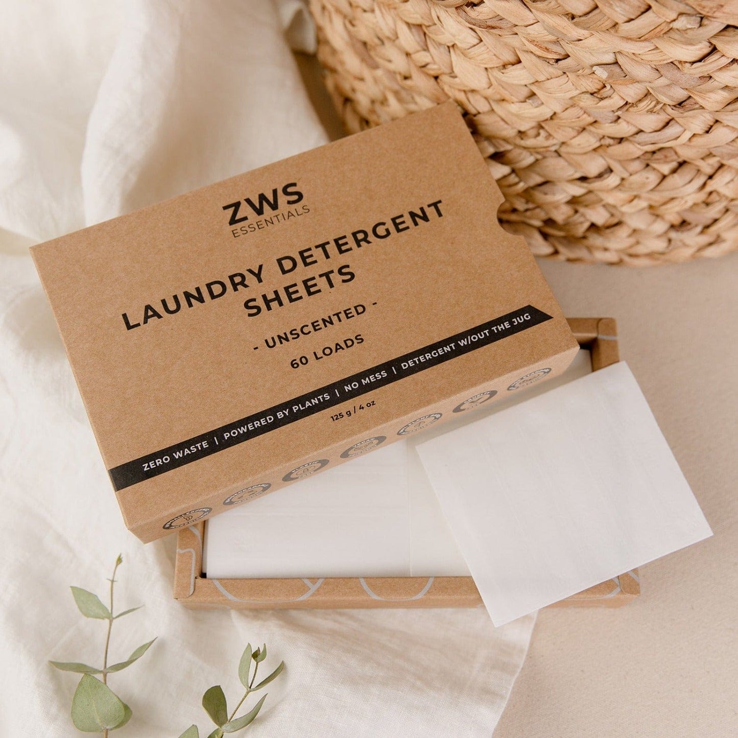 An open box of ZeroWasteStore.com Laundry Detergent Sheets sits on a beige surface, displaying the white sheets inside, with a woven basket, white cloth, and green leaves arranged nearby.