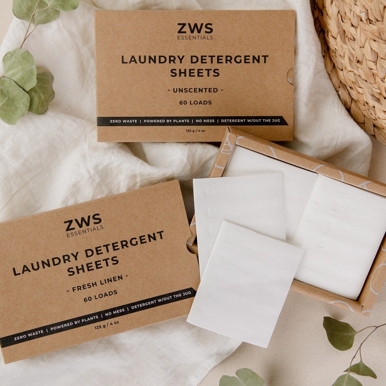 Two cardboard boxes of ZeroWasteStore.com Laundry Detergent Sheets are shown. One box is open, displaying the plant-based white sheets inside. Eucalyptus leaves and a woven basket are partially visible nearby.
