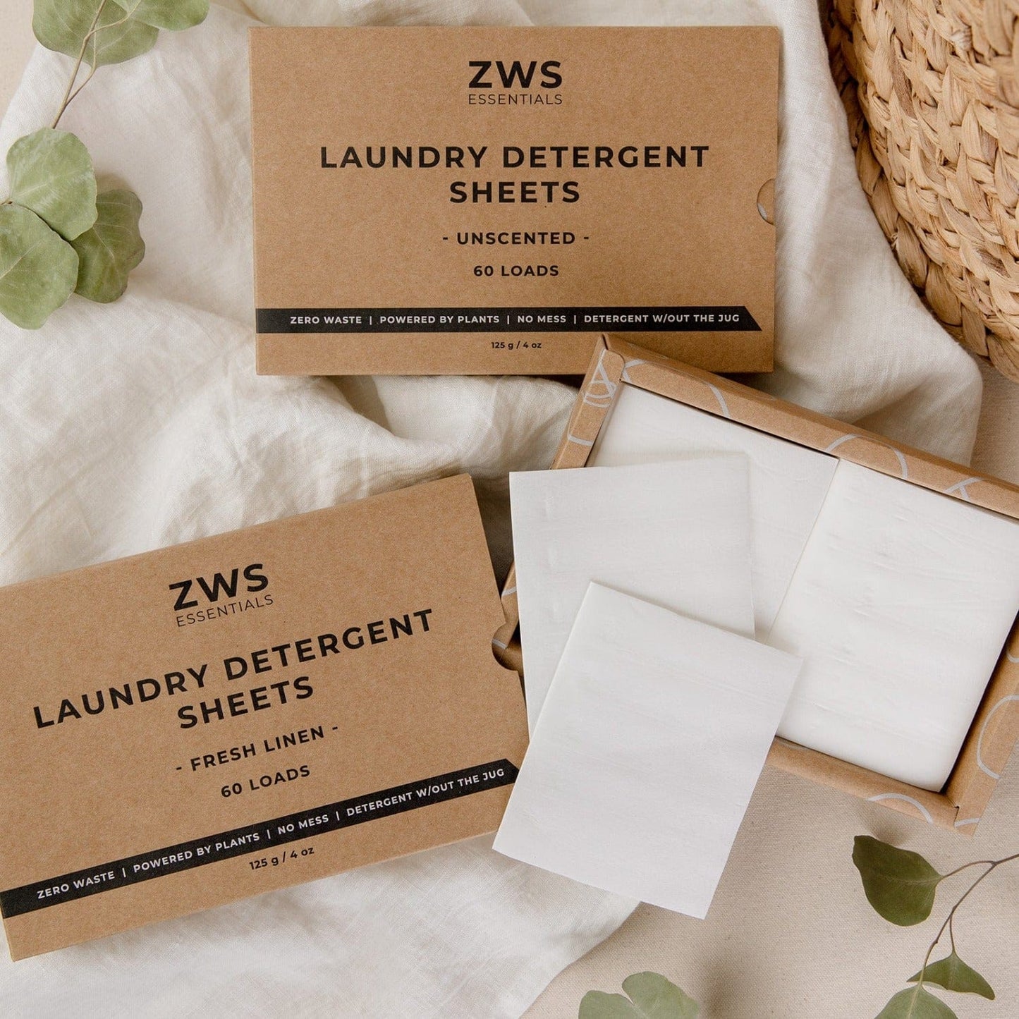 Two boxes of ZeroWasteStore.com Laundry Detergent Sheets—one Unscented, one Fresh Linen—are shown with two sheets outside the box, surrounded by fabric and greenery for a zero waste laundry solution.