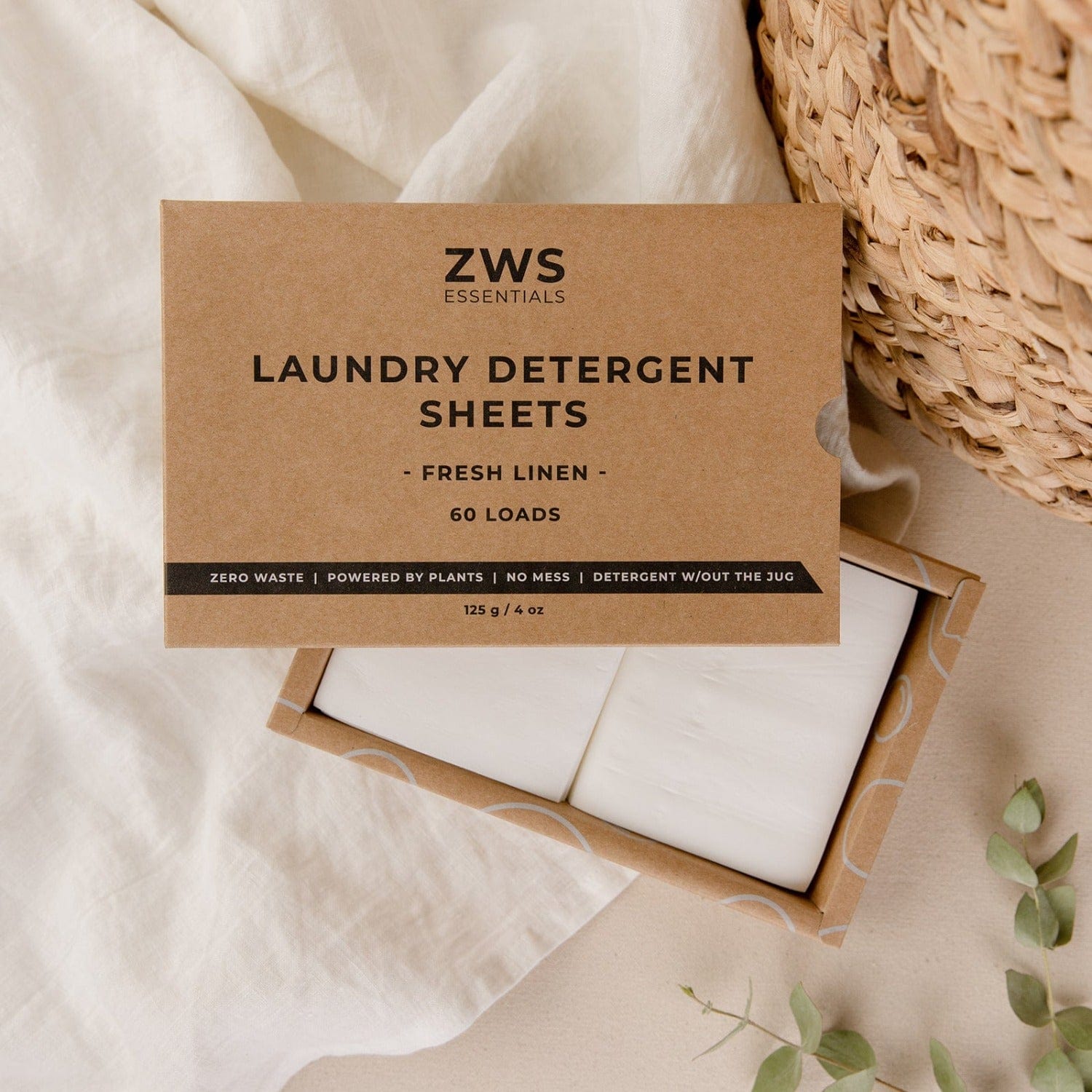 A box of ZeroWasteStore.com Laundry Detergent Sheets rests on cream fabric, with the white sheets visible inside. Nearby are a woven basket and a green plant, creating a fresh and eco-friendly vibe.