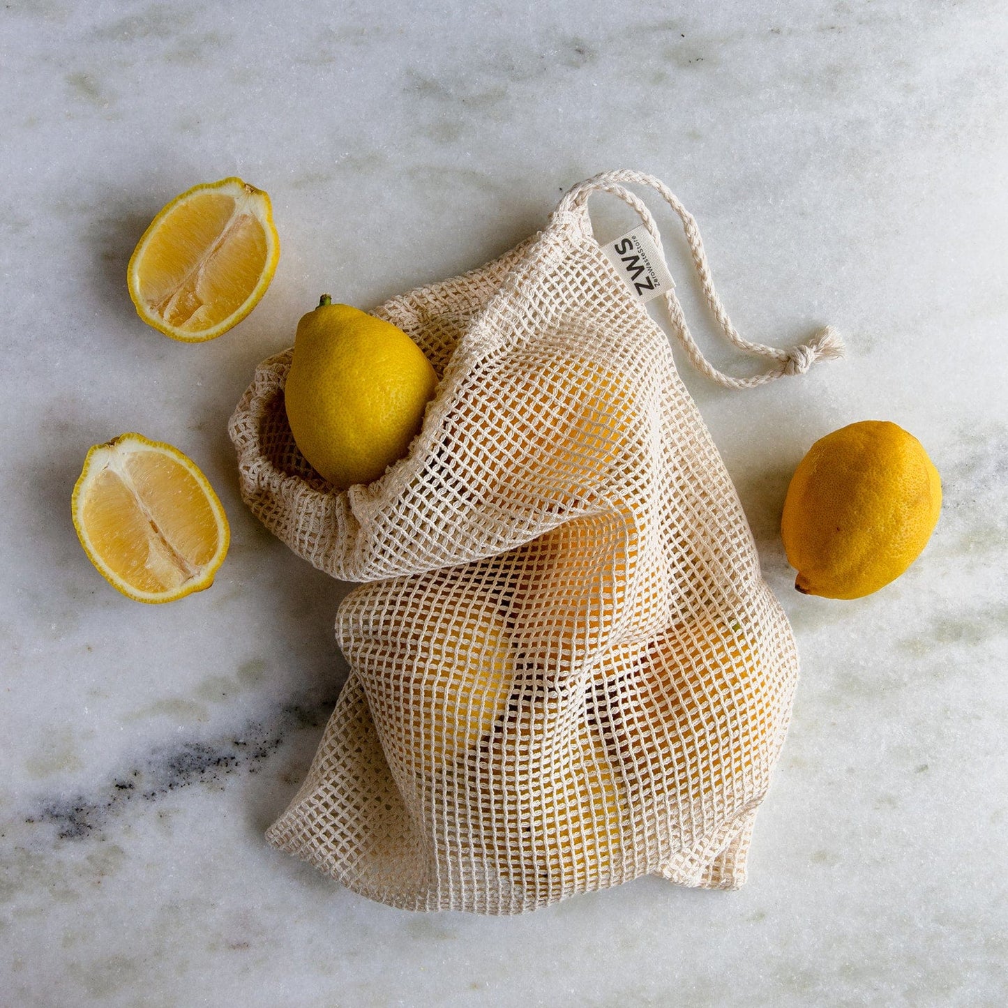 The ZeroWasteStore.com Mesh Produce Bag—available in multiple sizes—is made from organic cotton and pictured filled with lemons on a light marble surface, surrounded by whole and halved lemons for a perfect zero waste choice.