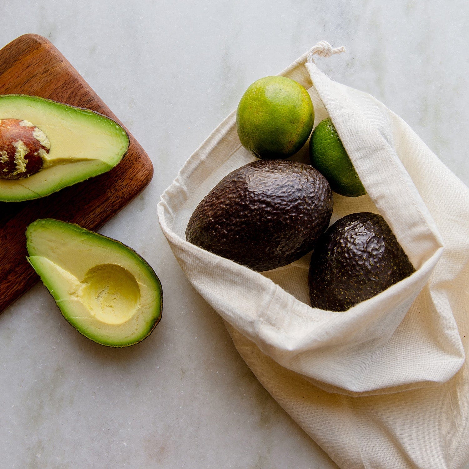 A ZeroWasteStore.com Muslin Produce Bag - Multiple Sizes holds two avocados and two limes on a marble surface beside a wooden board with a halved avocado, one half displaying the pit.