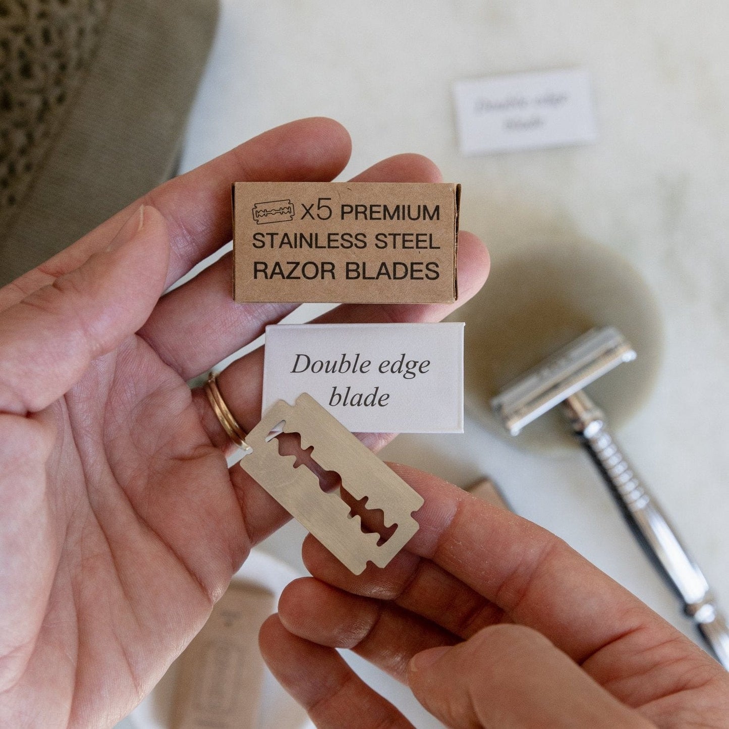 A person holds ZeroWasteStore.com Zero Waste Razor Replacement Blades (x5 Premium Stainless Steel) and a box, with a Zero Waste Razor on the countertop, highlighting an eco-friendly shaving routine.