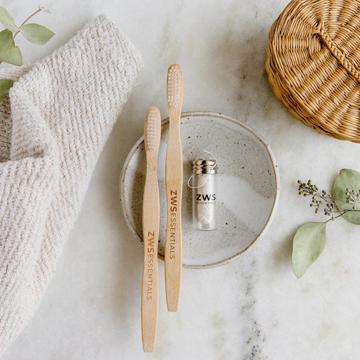 The Dental Care Mini Kit by ZeroWasteStore.com features two bamboo toothbrushes and a jar of biodegradable floss on a ceramic plate with a towel, wicker basket, and greenery—ideal for an elegant zero-waste dental routine.