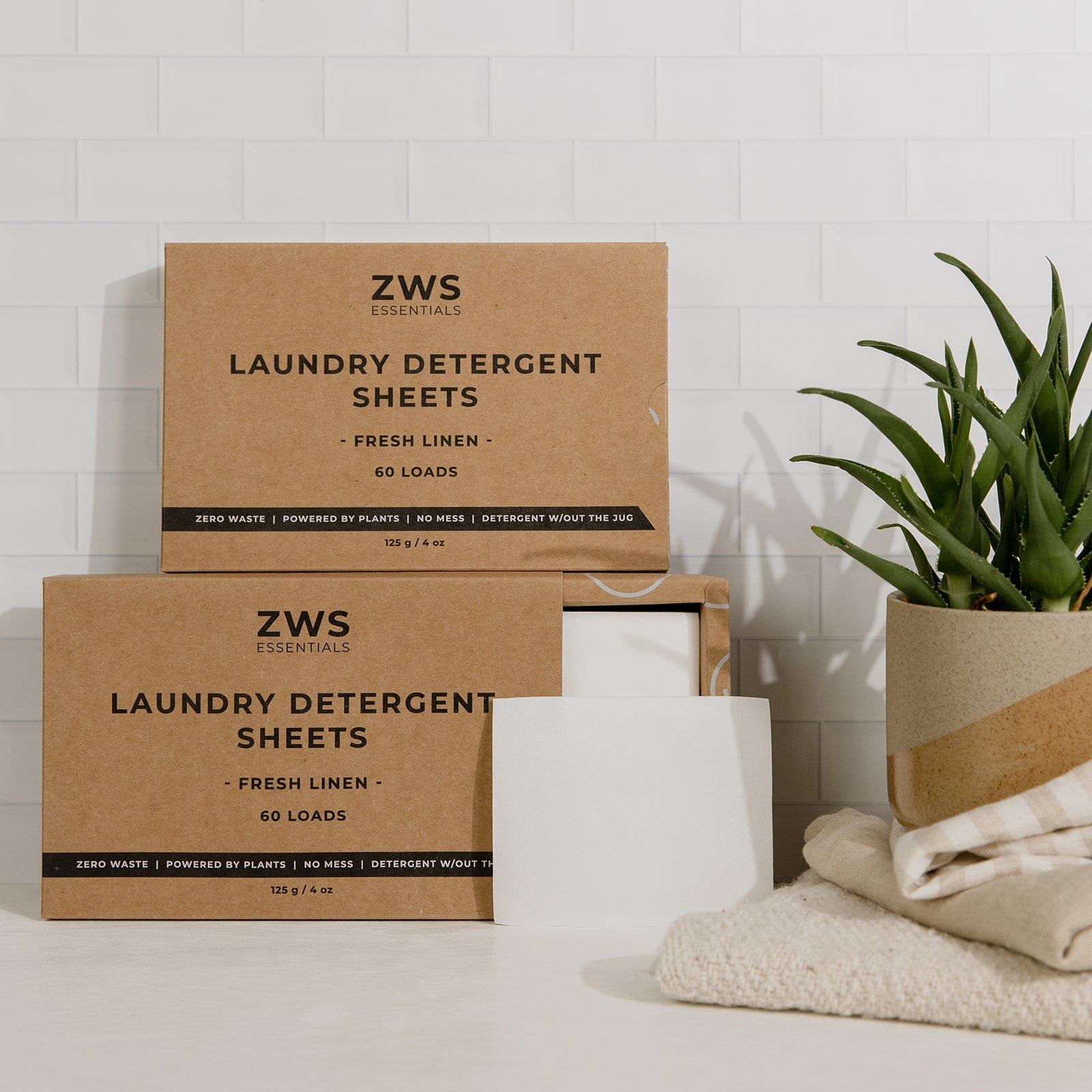 Two ZeroWasteStore.com Laundry Detergent Mini Kit boxes are displayed on a white surface next to a green potted plant, with a folded beige towel and one detergent sheet placed in front of the boxes.