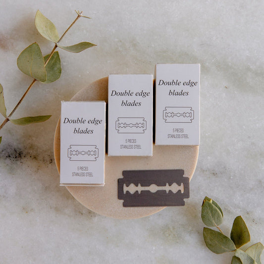 Three boxes of ZeroWasteStore.com’s Zero Waste Razor Replacement Blades and a single blade rest on a dish, with green eucalyptus leaves on marble—an eco-friendly addition to your sustainable shaving routine.